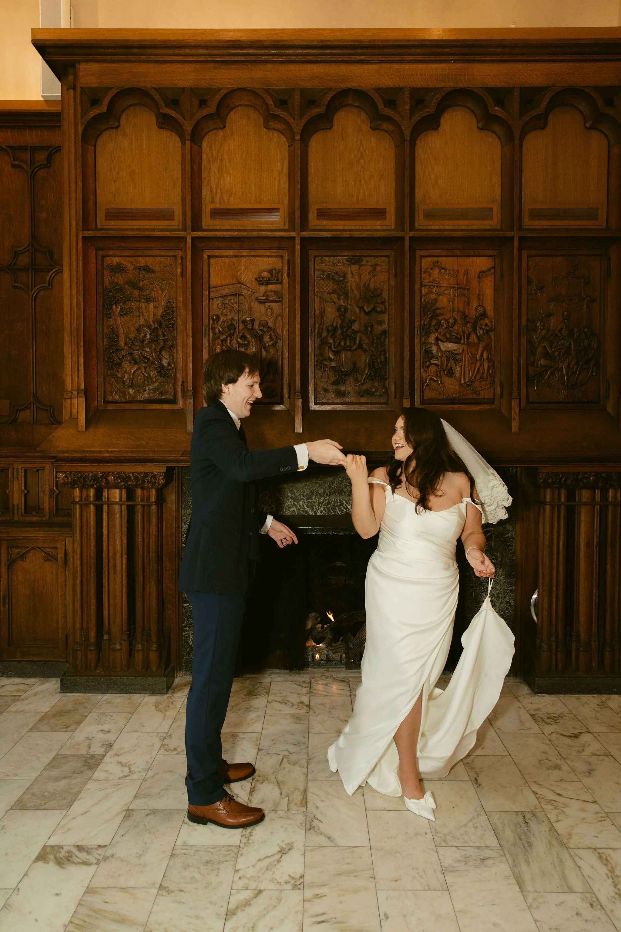 A bride and groom are dancing in front of a wooden mantel with carved panels in an elegant interior. The groom is wearing a dark suit and brown shoes, and the bride is in a white dress with a veil, smiling as they hold hands.