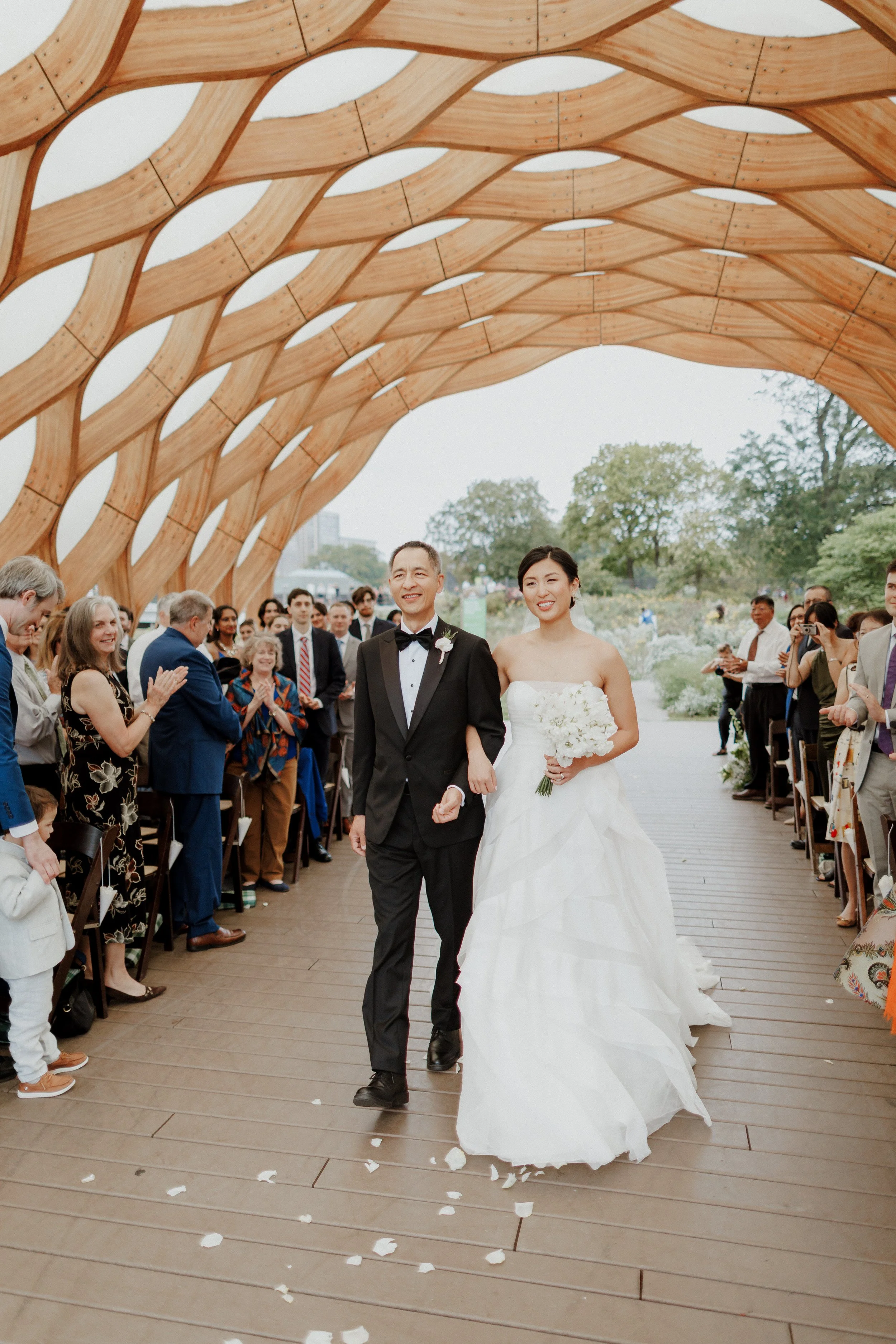 A bride and groom walking hand in hand down an aisle at their wedding, surrounded by family and friends in an outdoor setting with a wooden arch overhead and trees in the background.