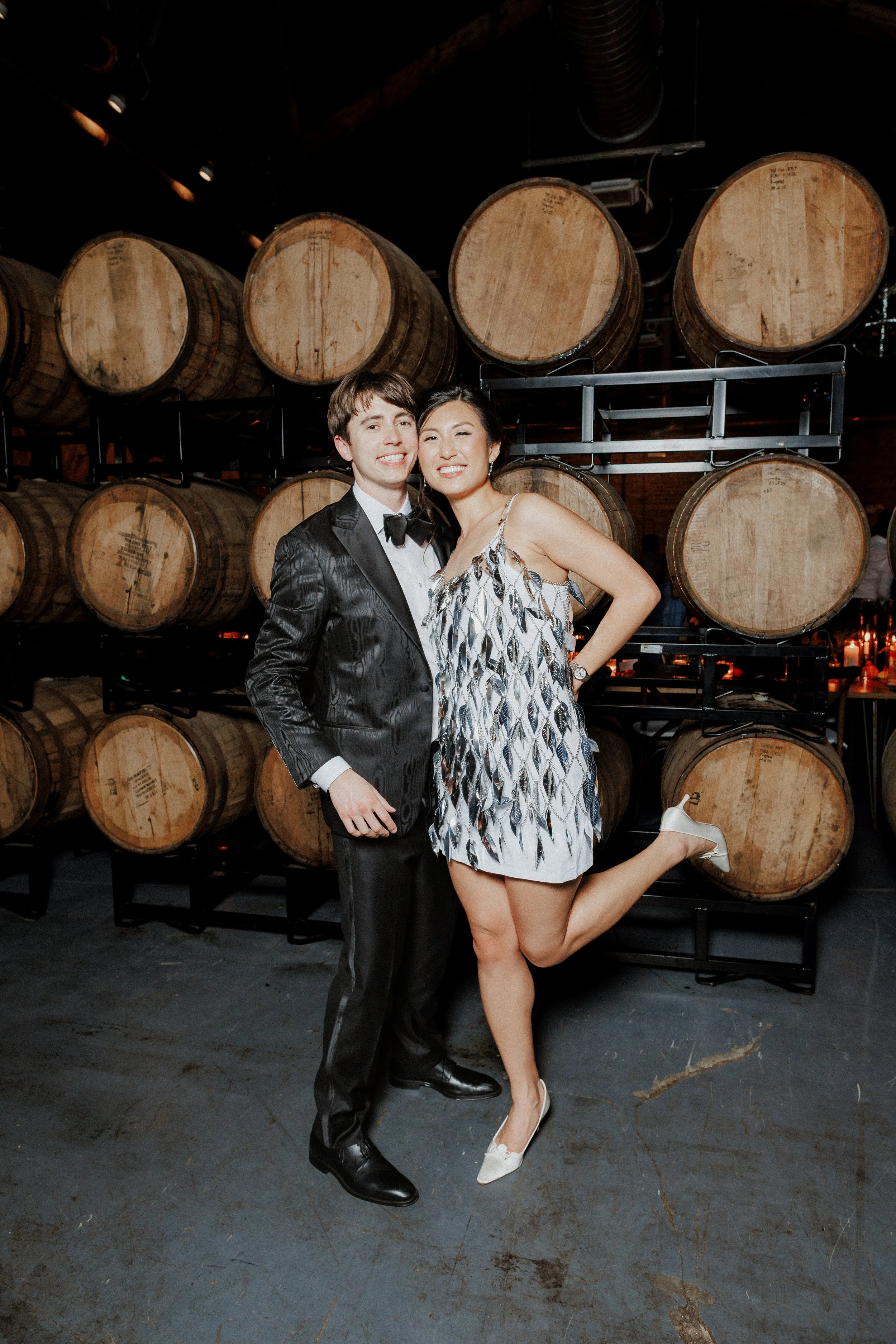 A man in a tuxedo and a woman in a dress with high heels pose together in a barrel warehouse, with the woman on one foot and the man smiling.