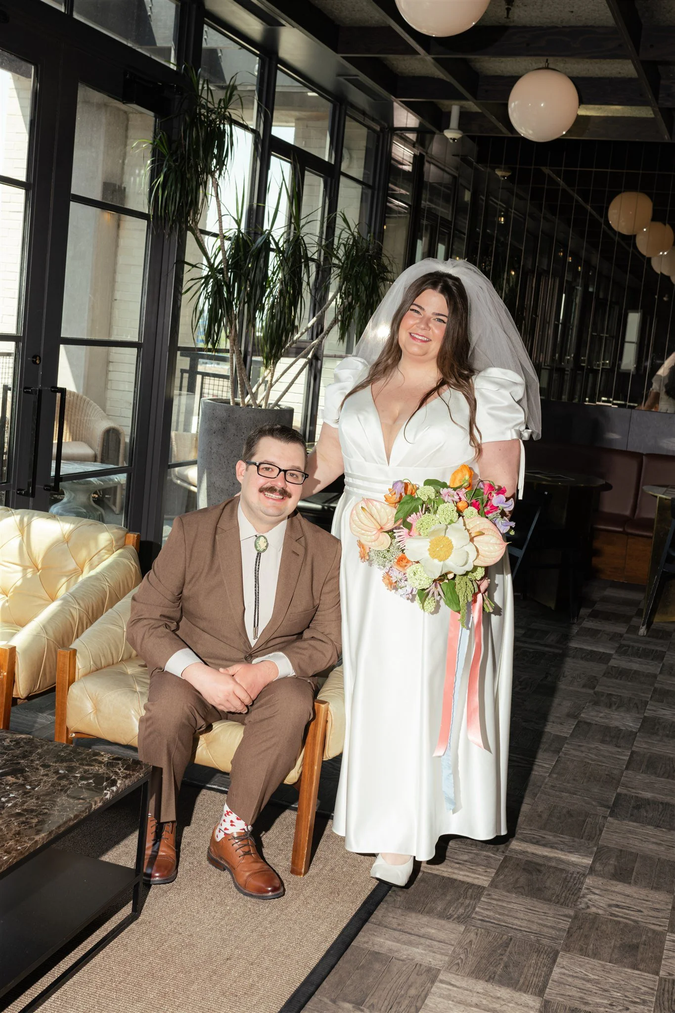 A woman in a white dress and veil holding a colorful bouquet stands beside a man in a brown suit. They are in a modern indoor setting with large windows, potted plants, and round hanging lights.