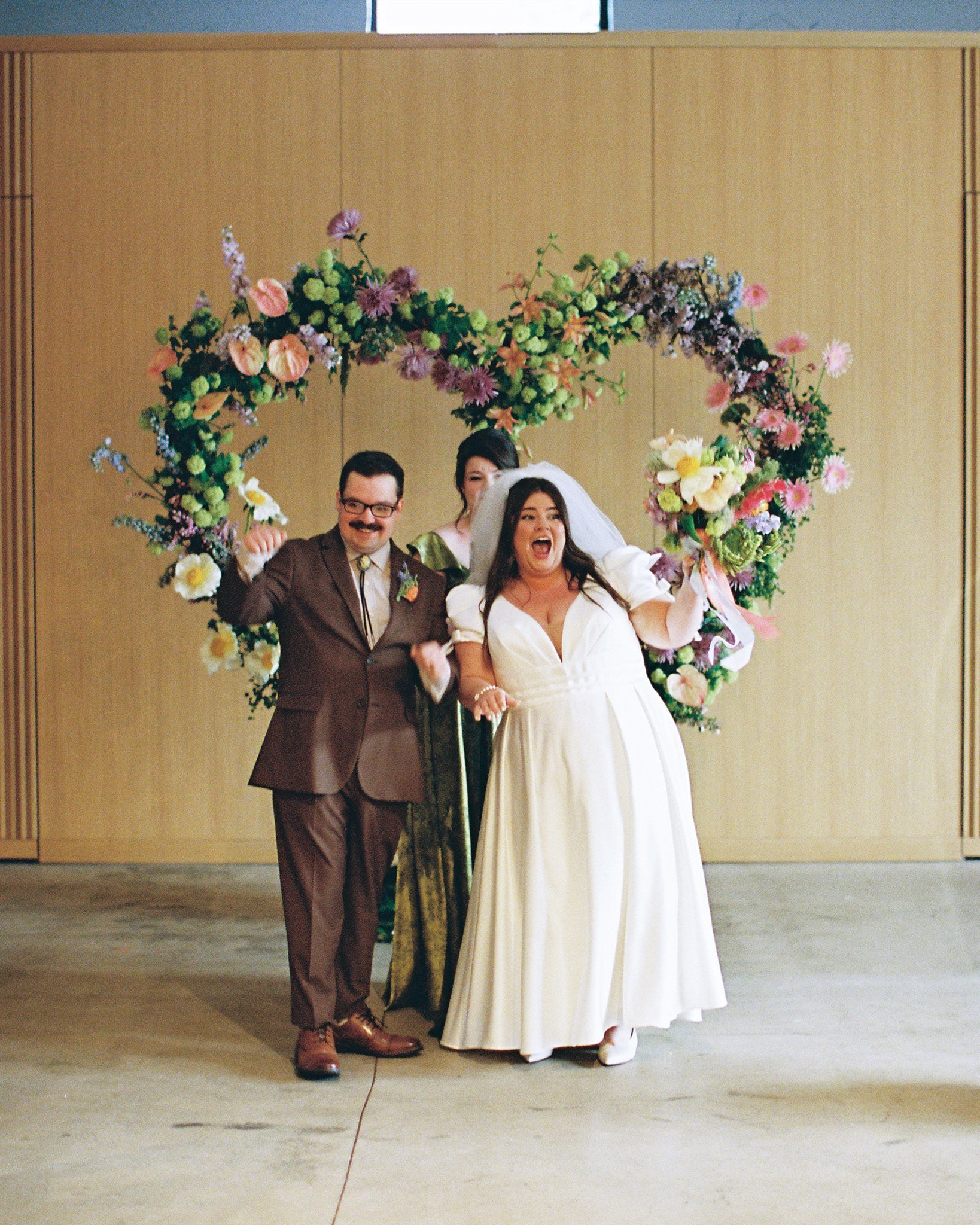 A bride and groom celebrating their wedding reception, standing under a floral arch. The groom is wearing a dark brown suit and glasses, and the bride is in a white wedding dress and veil, smiling happily. An officiant or another person stands behind
