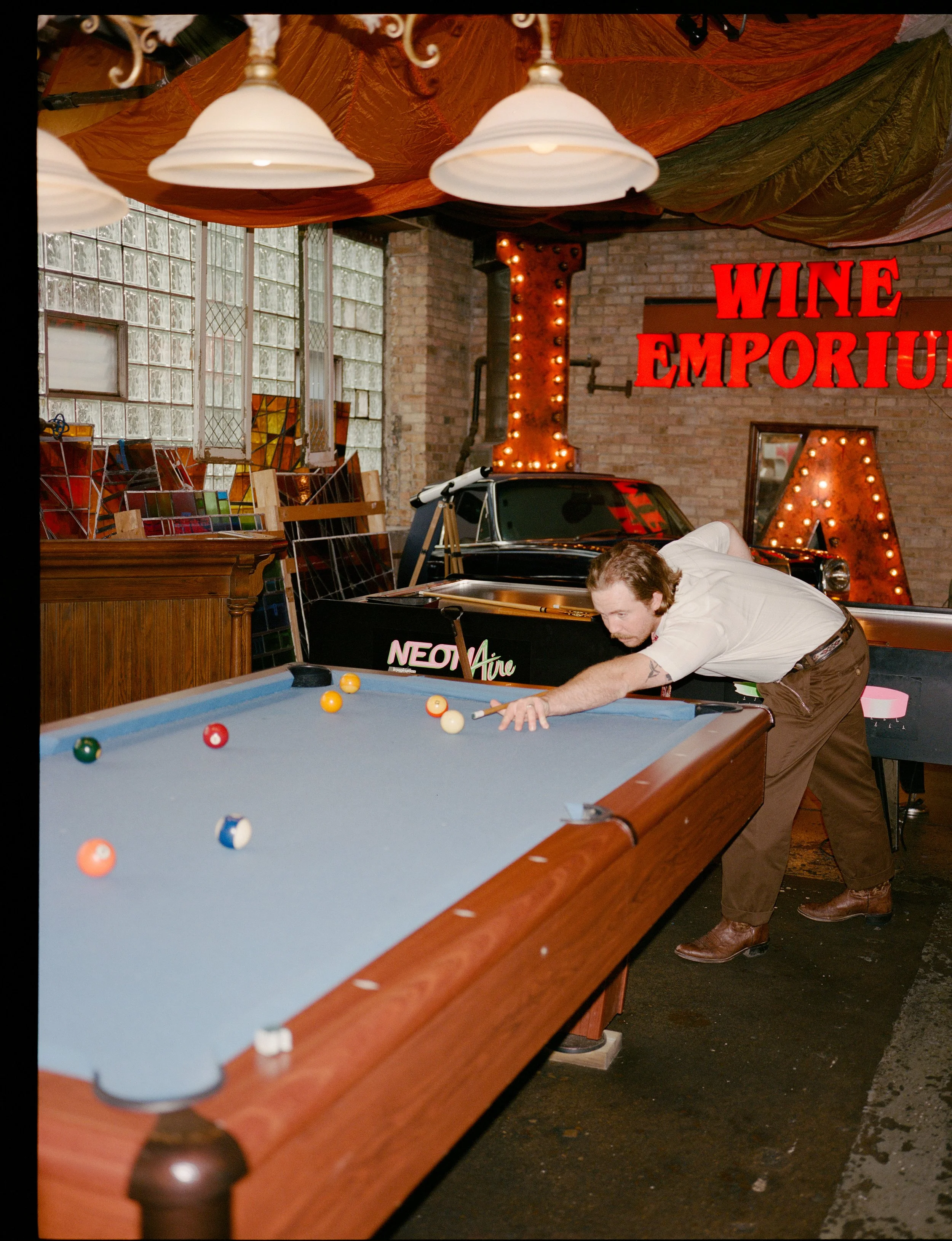 A man with long hair and tattoos wearing a white shirt and brown pants playing pool on a blue-felt table in a room with brick walls and large glass block windows. There are various artworks and sculptures on shelves and walls, and large illuminated s