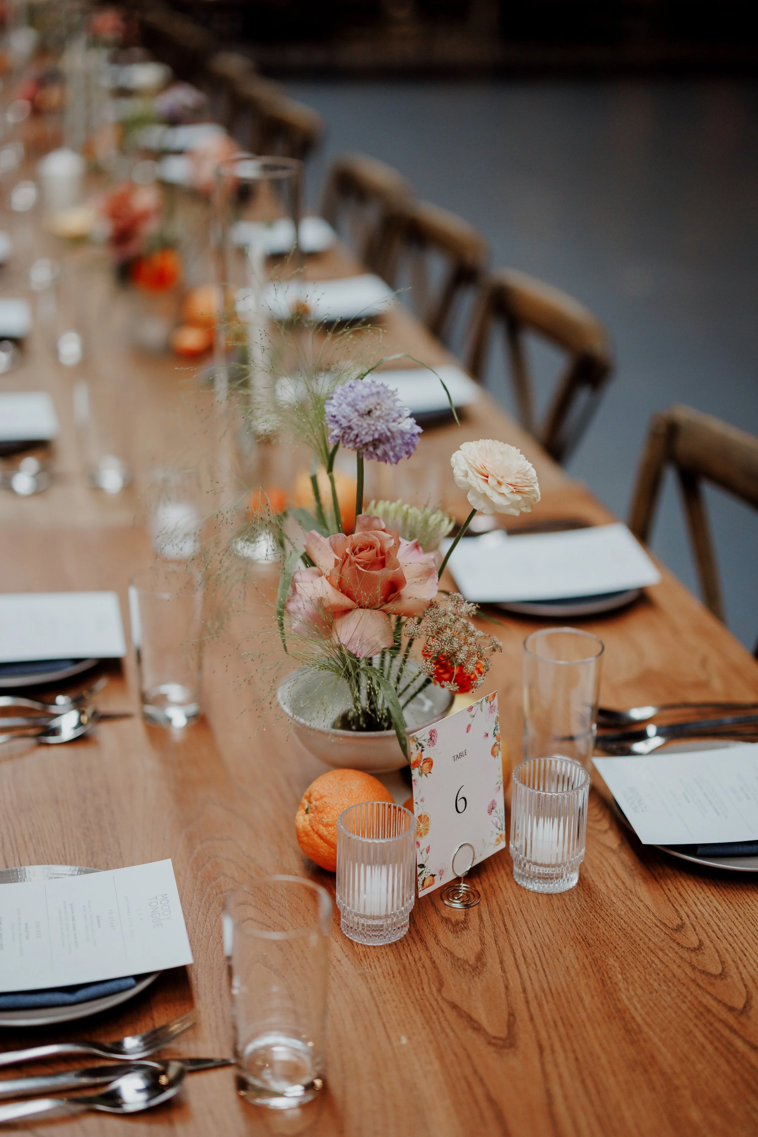 A long wooden dining table set for a meal with chairs on either side, decorated with flower arrangements, clear glasses, white napkins, and small menu cards.