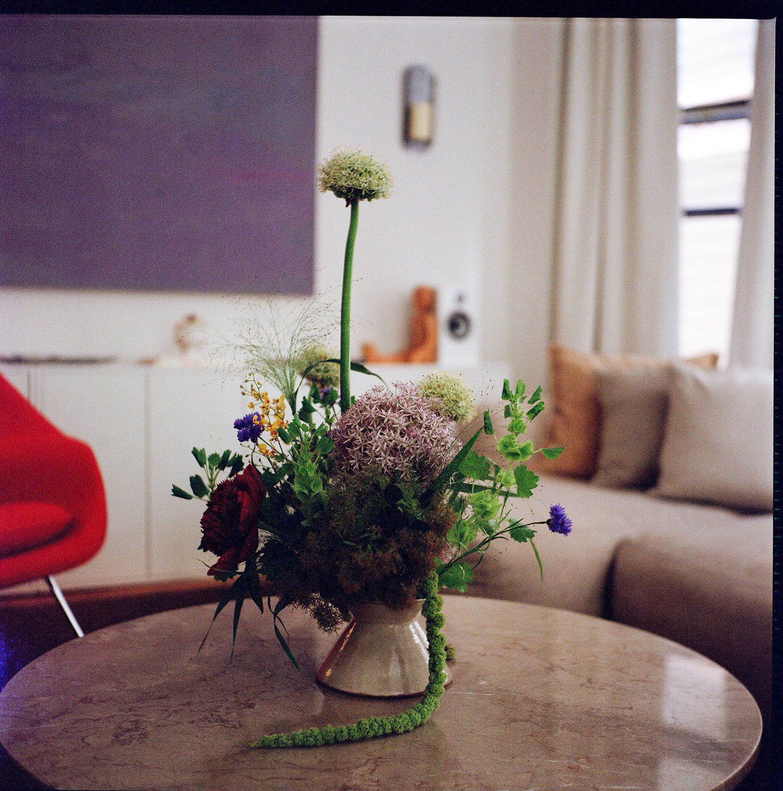 A flower arrangement with various colorful flowers in a ceramic vase on a round wooden table in a living room with beige and brown furniture and white curtains.