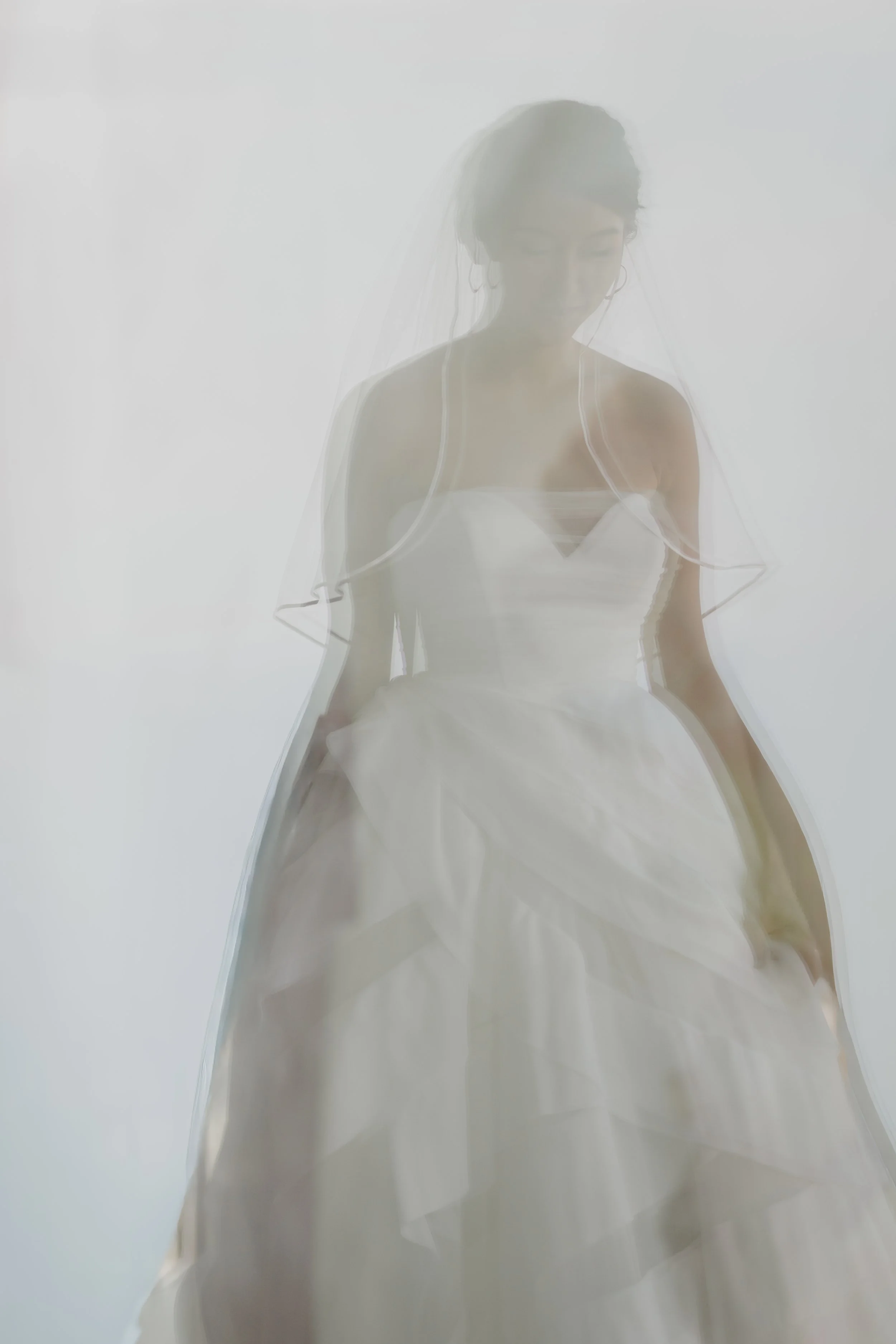 A bride wearing a wedding gown and veil with a sheer overlay, standing with her head slightly bowed and eyes closed, in front of a plain white background.