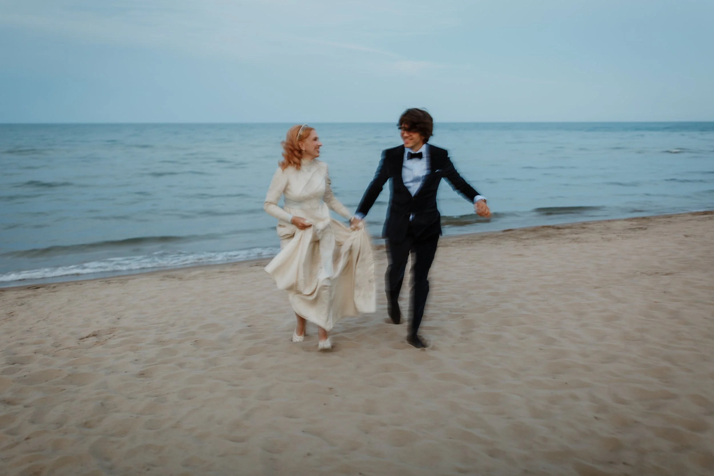 A woman in a long white dress and a man in a tuxedo holding hands and walking on the beach near the ocean, smiling and laughing.