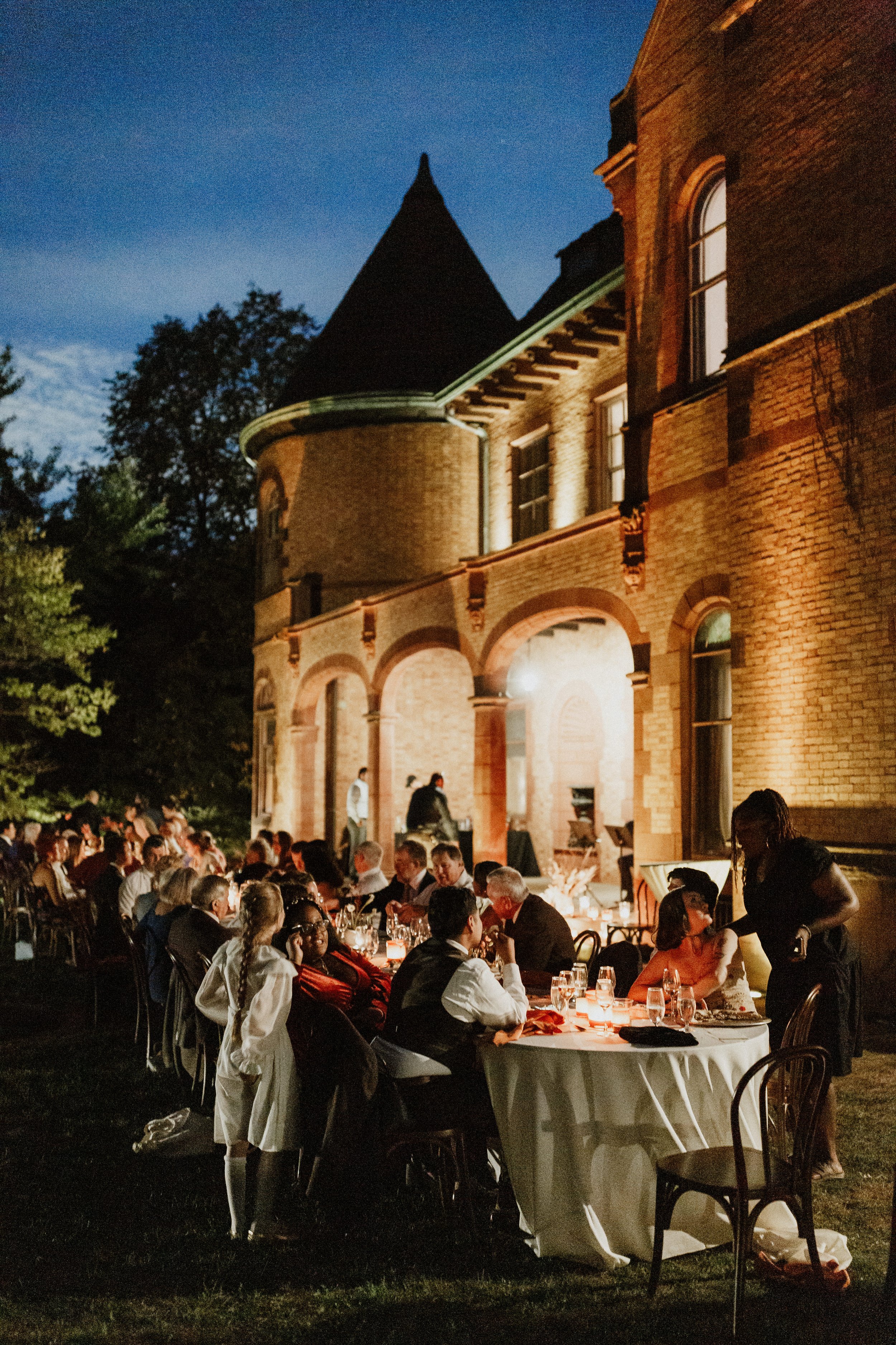 Nighttime outdoor banquet with long tables, white tablecloths, candles, and guests dining in front of a historic brick building with arched windows and turret, surrounded by trees.