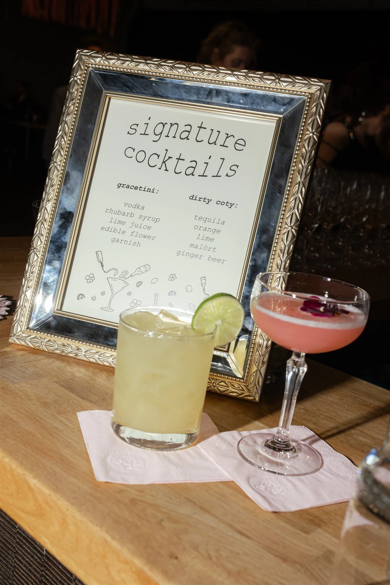 A framed sign lists signature cocktails with ingredients, placed on a table with two cocktails in front, one with lime and the other pink, in a dimly lit setting.