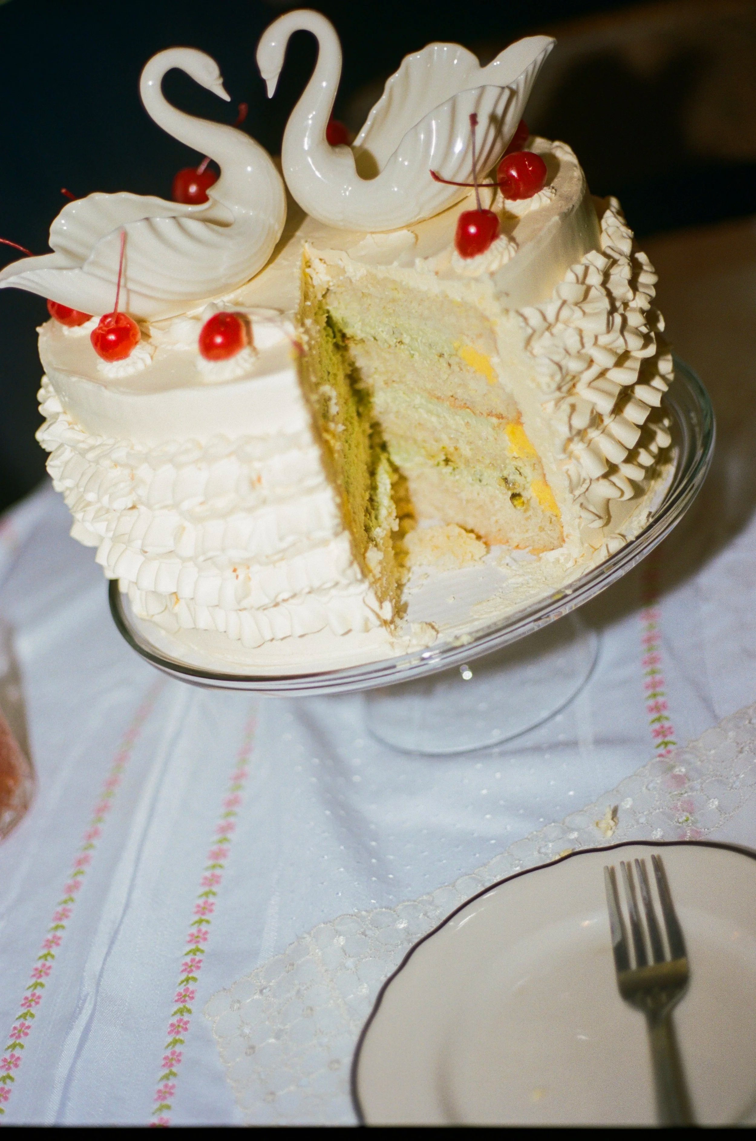 A slice of white cake with white frosting and red cherries on top, decorated with swan-shaped white meringue and piped frosting, served on a glass cake stand.