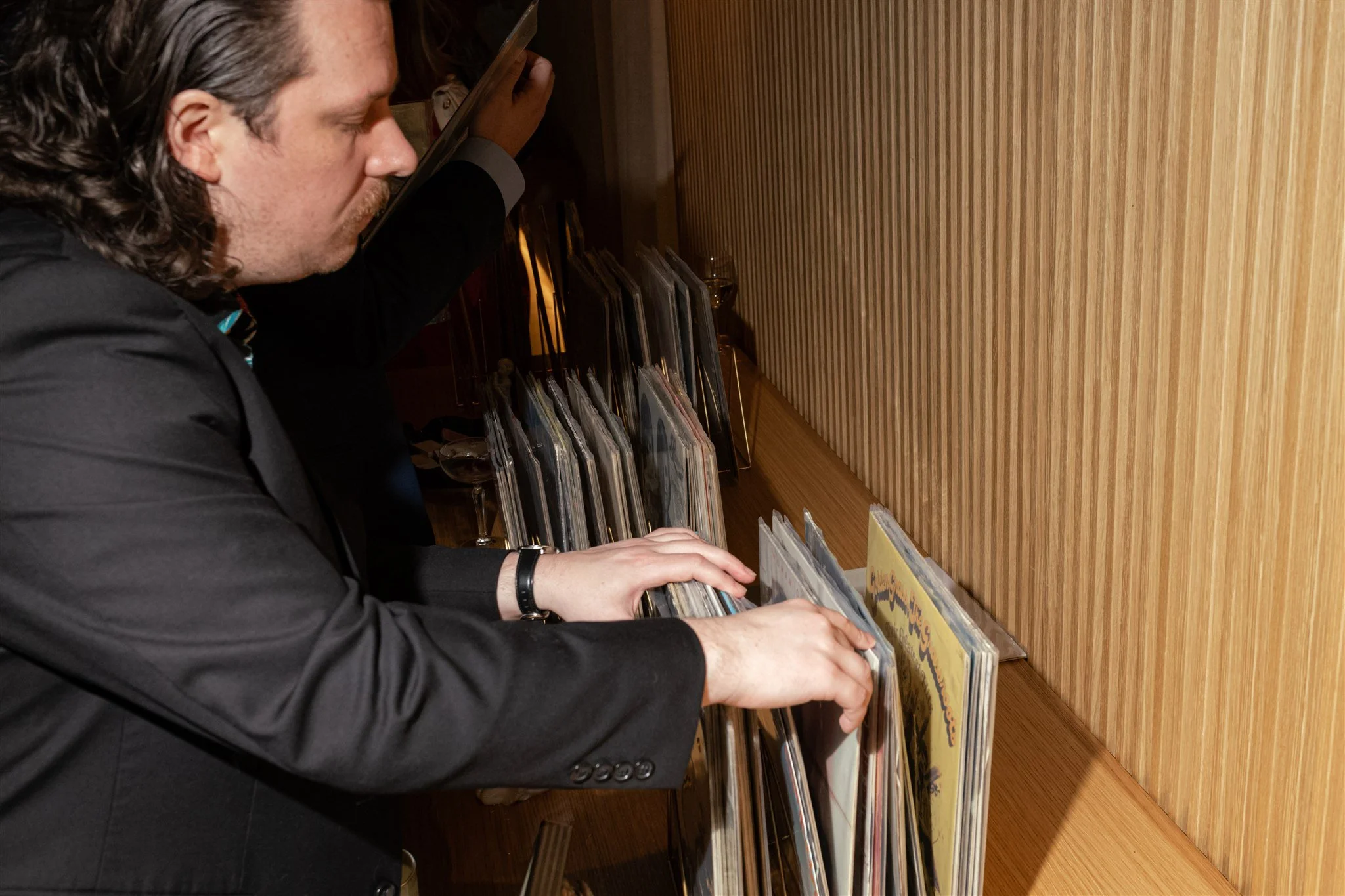 A man with long dark hair and a beard wearing a black suit is browsing vinyl records in a store with wood-paneled walls.