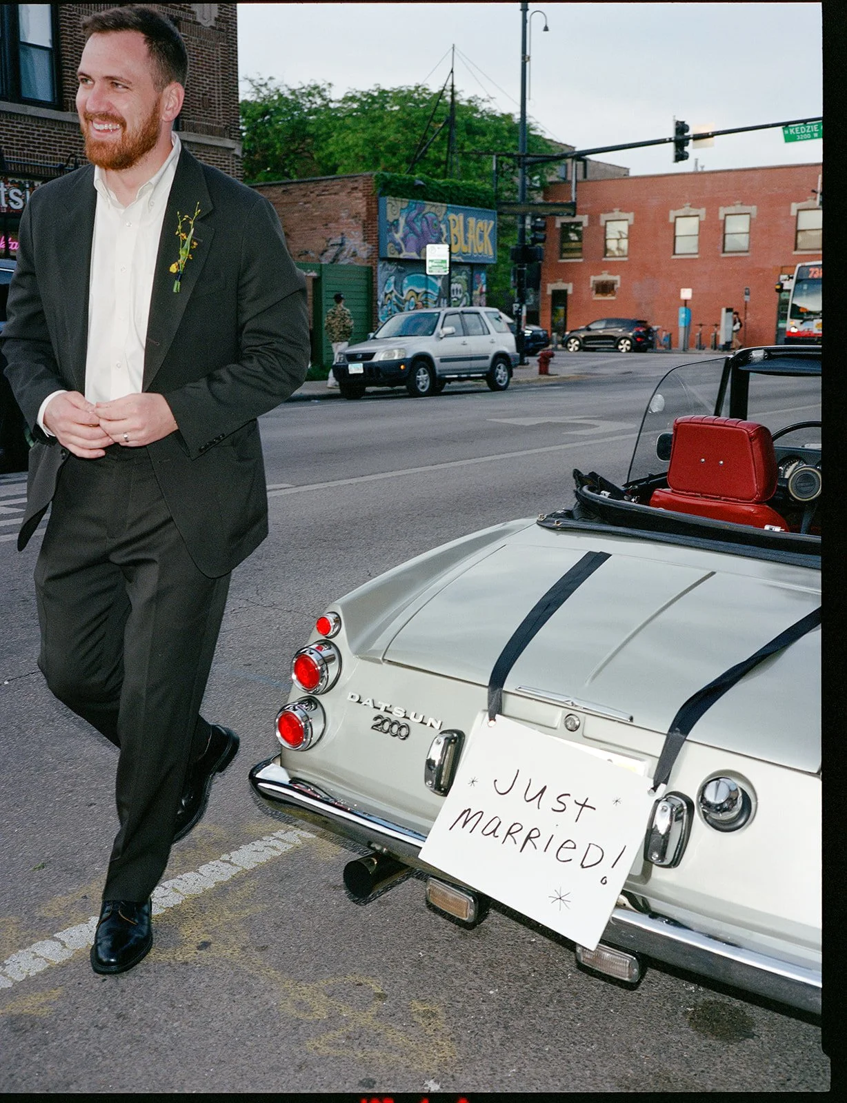 A man in a black suit and white shirt standing next to a vintage white Datsun 2000 convertible with a sign that reads 'Just Married' hanging on the back. The man is smiling and holding his hands together.