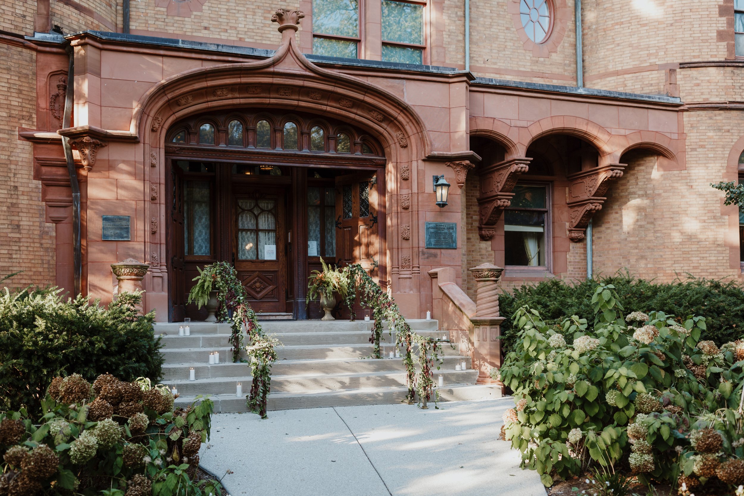 Entrance of a historic brick building decorated with floral arrangements and candles for a wedding, featuring steps leading up to a wooden door and surrounded by lush greenery.