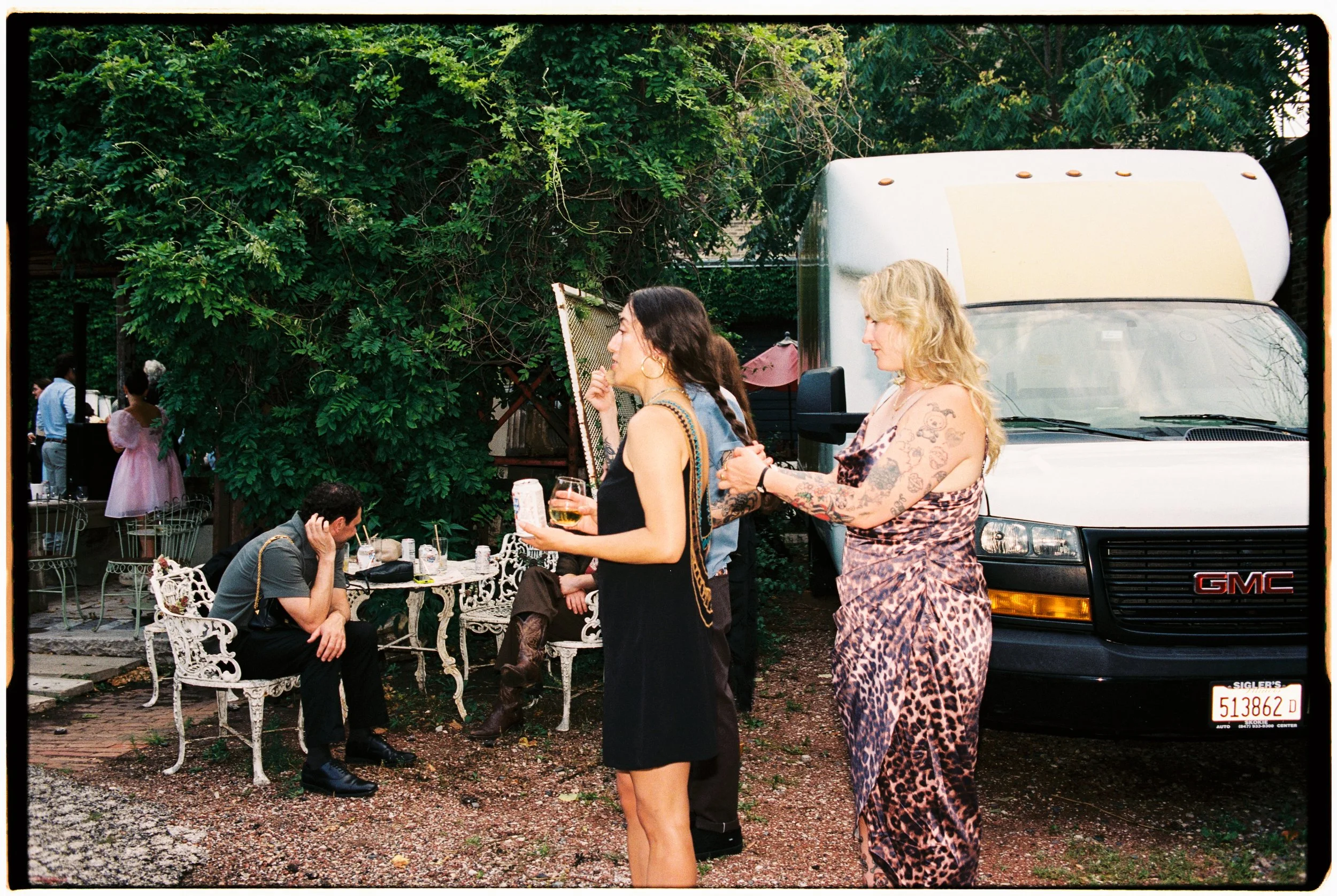 People gathered outdoors, some standing and some seated on white ornate metal chairs, in a garden area with lush green trees in the background. Two women are in the foreground, one with dark hair wearing a black dress and holding a drink, the other w