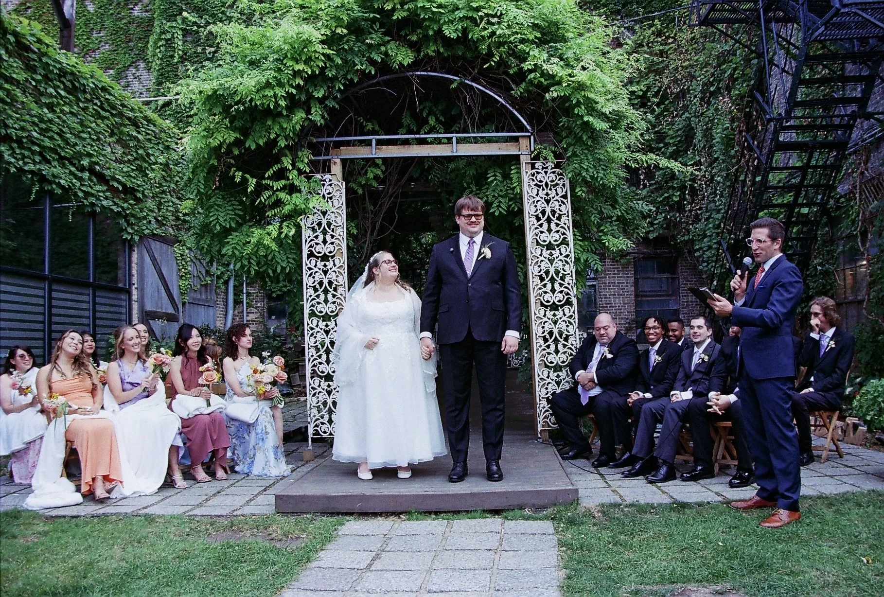 A wedding ceremony outdoors with the bride and groom holding hands, surrounded by friends and family sitting and standing on a brick patio with lush greenery and trees in the background. A man in a suit is reading vows with a microphone.