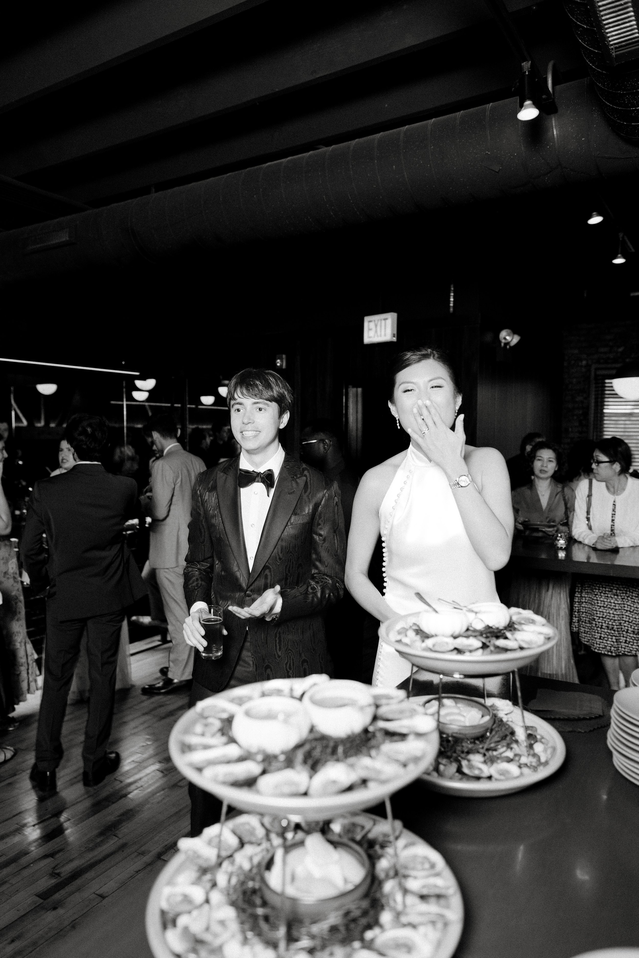 A black and white photo at a social gathering with people dressed formally. In the foreground, there is a tiered tray with assorted finger foods and snacks. Two individuals, a man in a tuxedo with a bow tie and a woman in an elegant dress, are promin