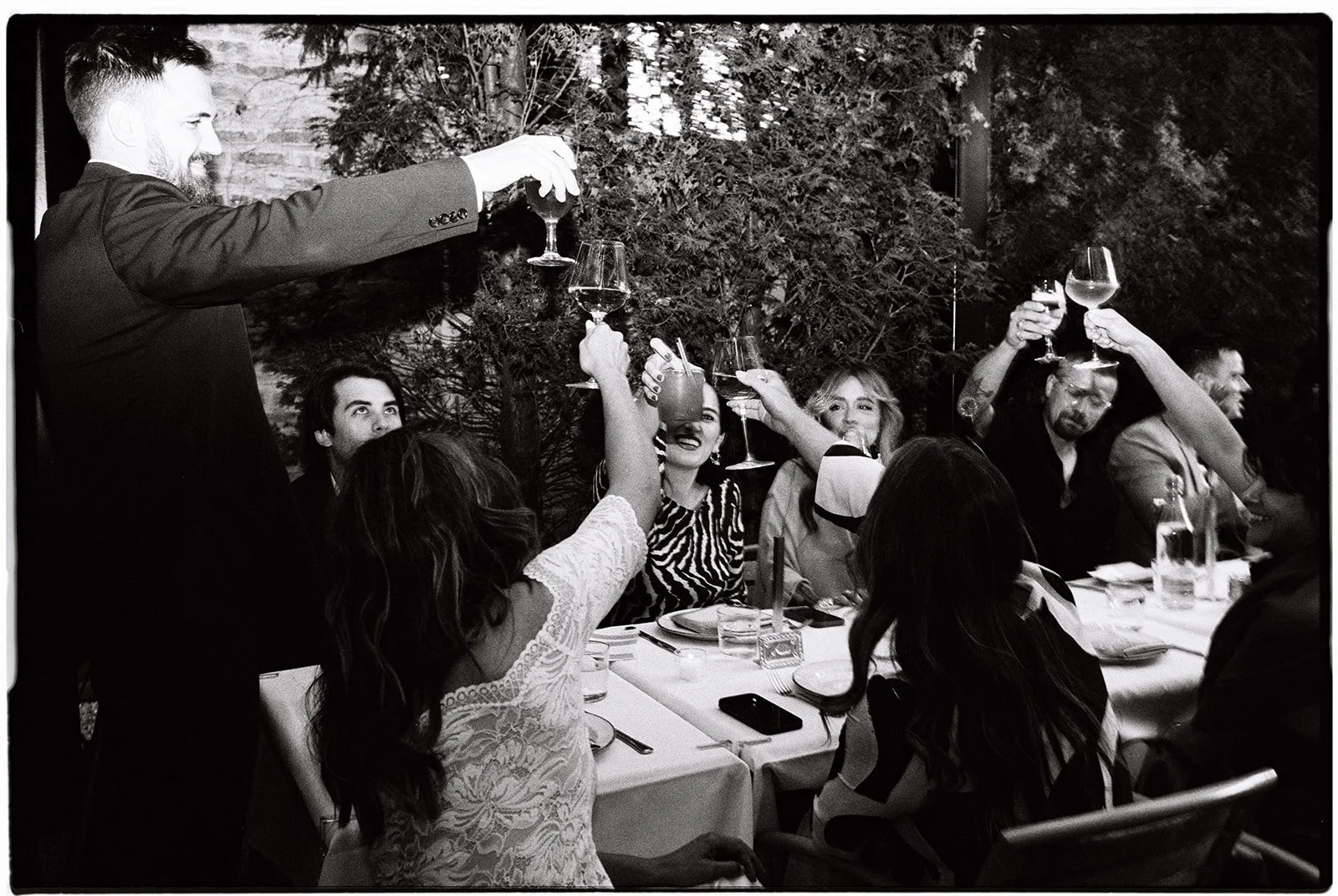 Group of people at dinner party raising wine glasses in a toast, with smiling faces, seated at a dining table outdoors surrounded by trees.