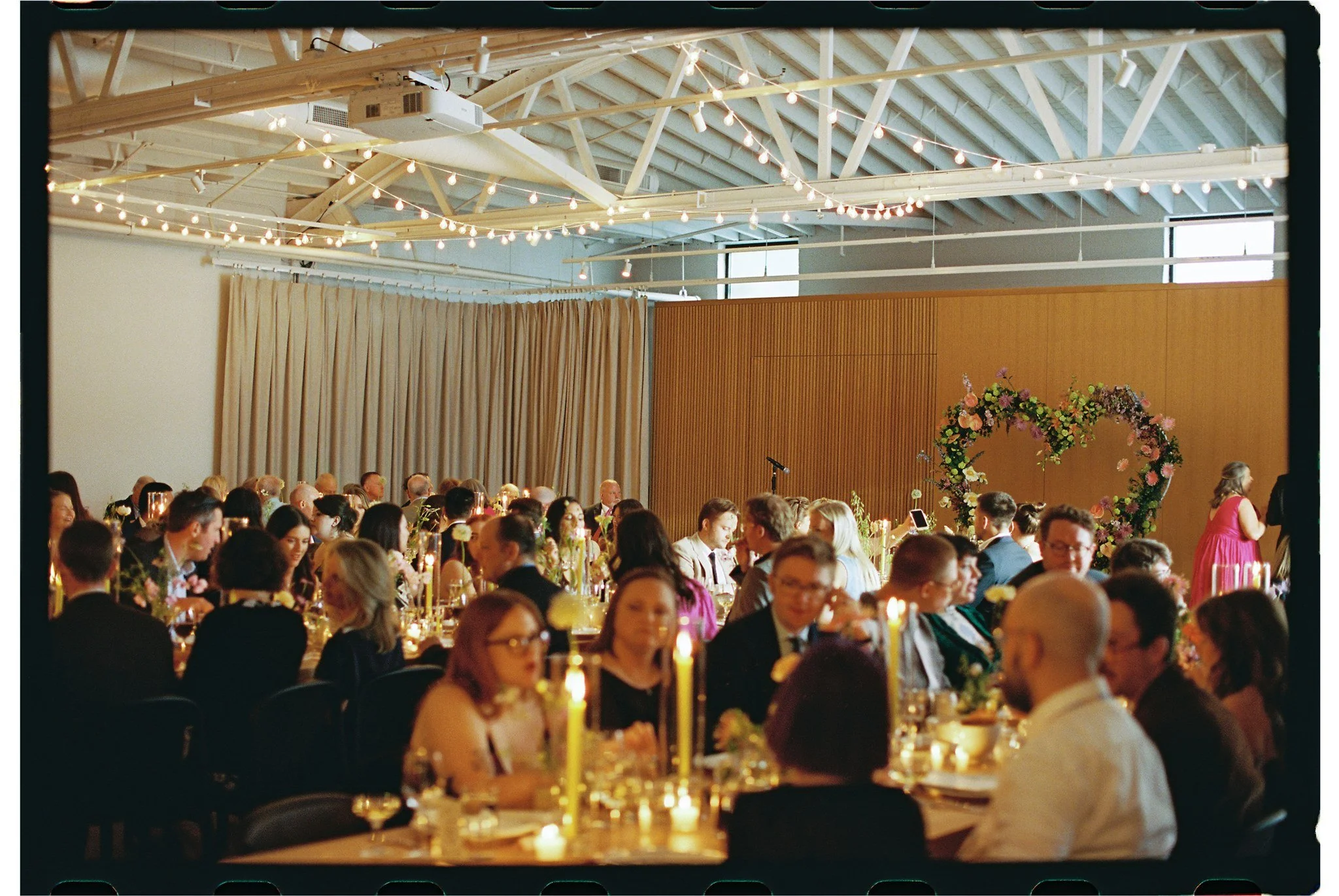 At a formal event or wedding reception, numerous guests are seated at round tables decorated with floral centerpieces and lit candles, with a large floral arch in the background and string lights hanging from the ceiling.