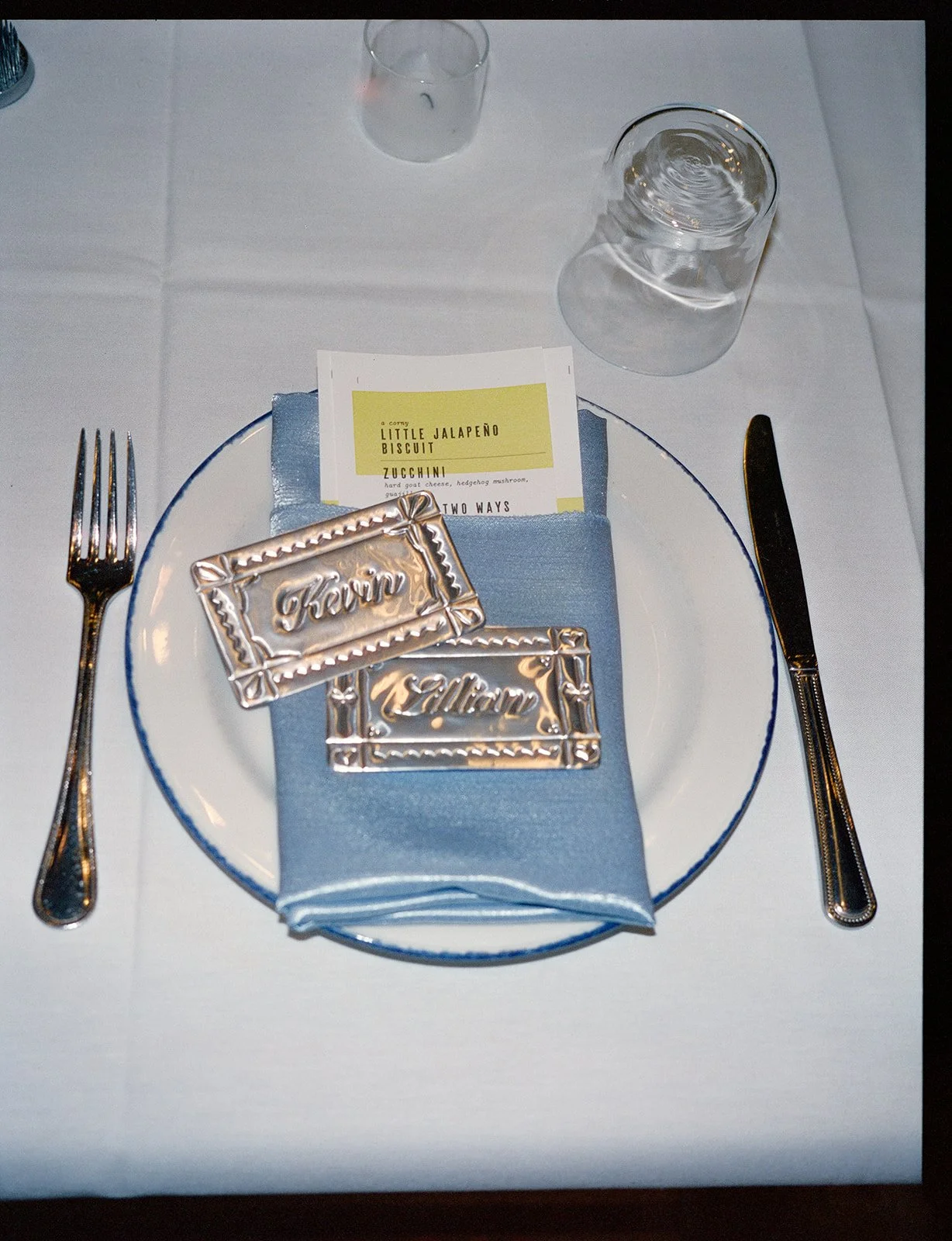 Place setting with a white plate and blue napkin, silver fork and knife, two silver chocolate bars with cursive writing, a menu card, and two empty glasses on a white tablecloth.