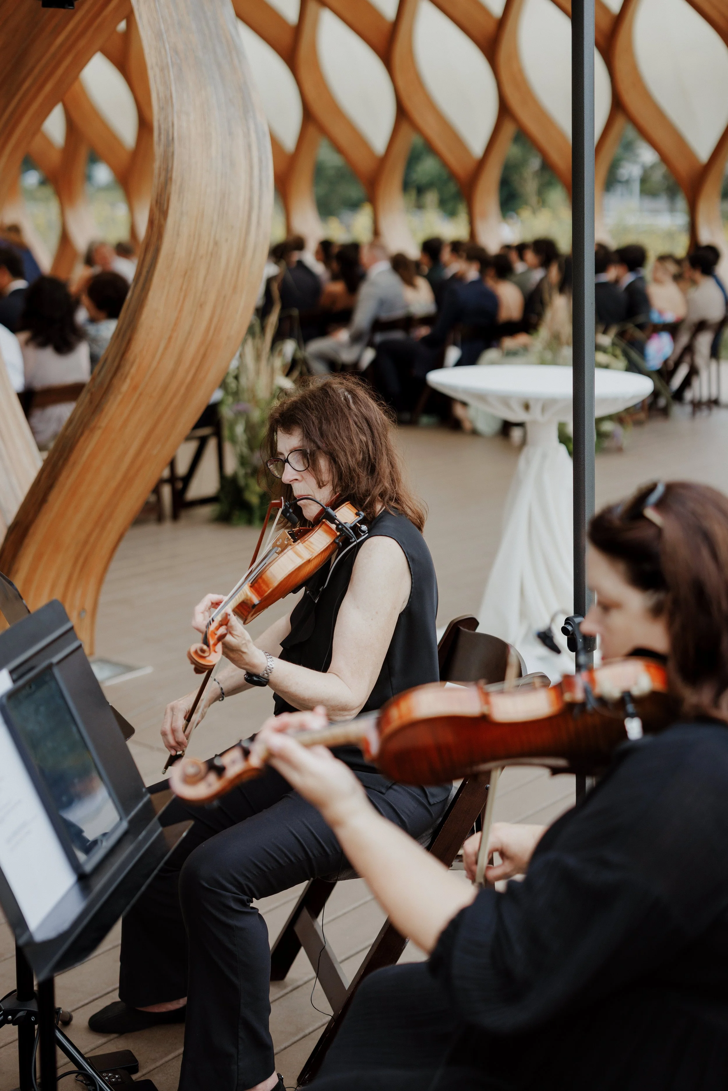 Two women playing violins in an outdoor setting, with many seated people in the background, under a wooden architectural structure.