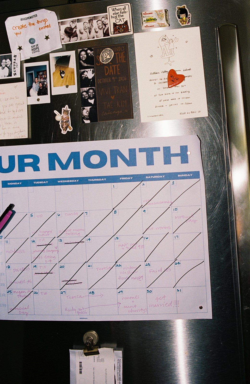 A refrigerator door with various magnets, photos, and notes, including a calendar page for the month with handwritten events, a black chalkboard-style card about a date, and small decorative items.