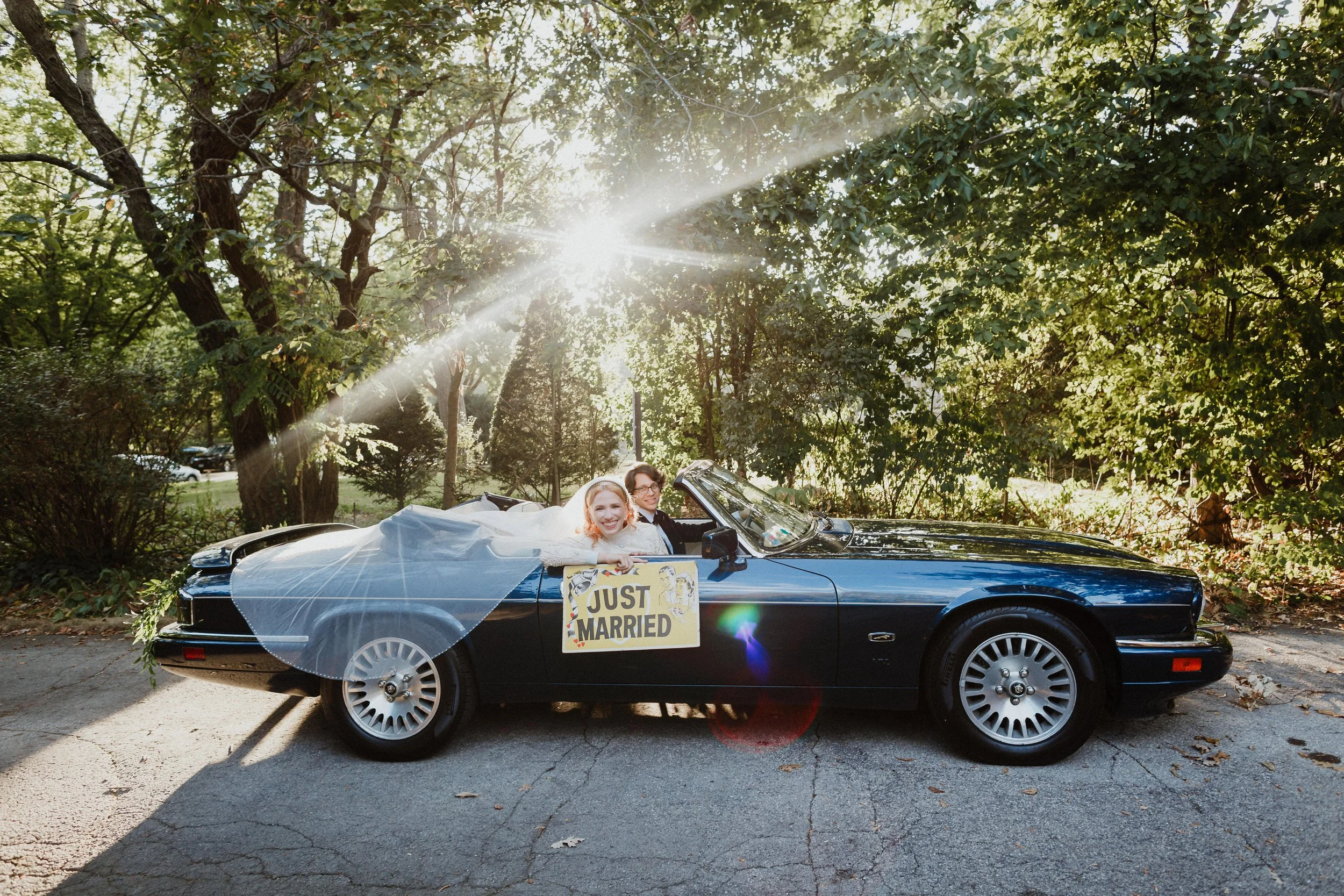 A newlywed couple sitting in a black convertible car decorated with a white veil and a 'Just Married' sign, parked under tall green trees with sunlight filtering through the leaves.