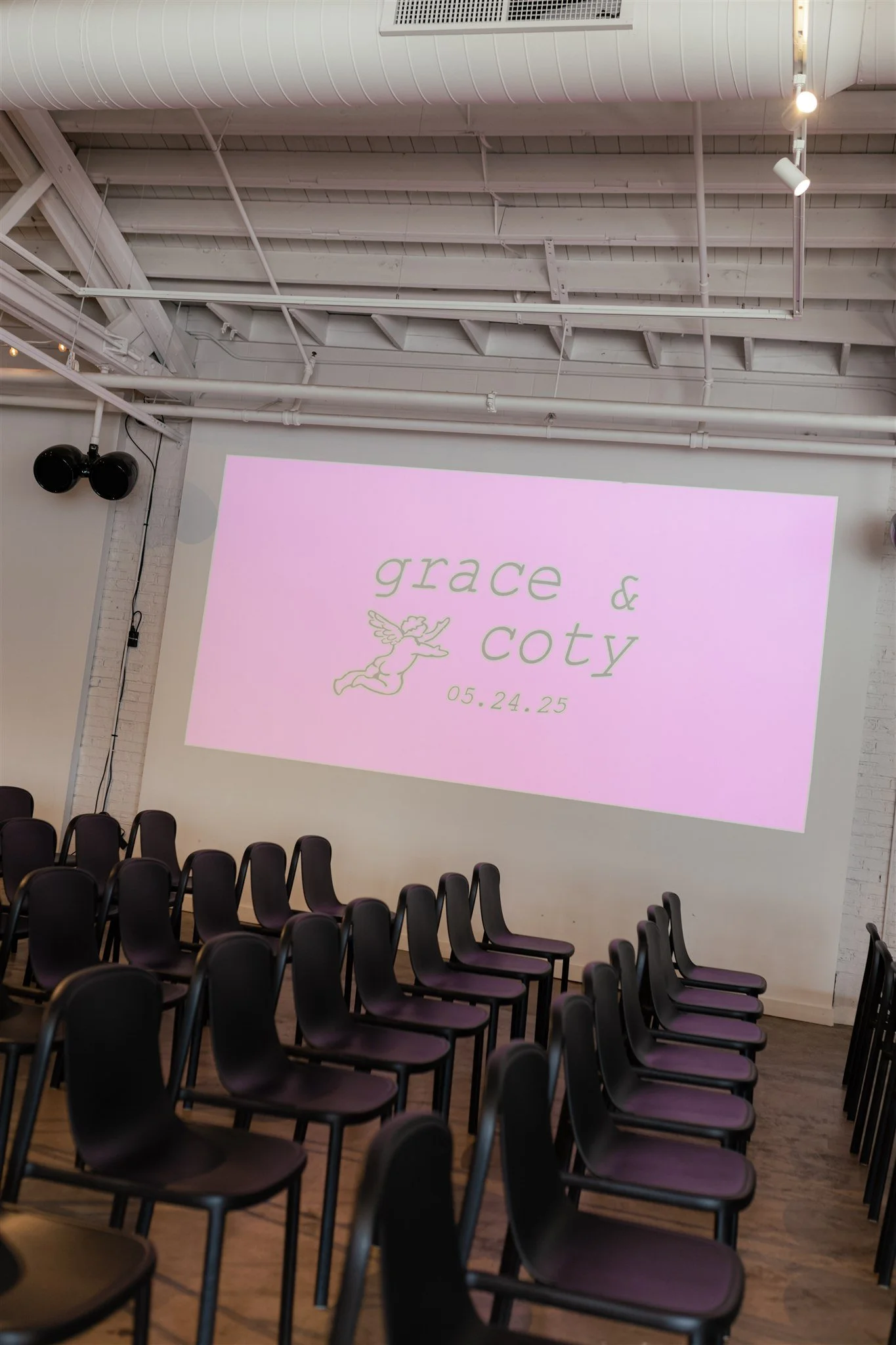 A large room with rows of black chairs arranged facing a wall with a pink projected screen displaying the words 'grace & coty' and a angel drawing, along with date '05.24.25'.