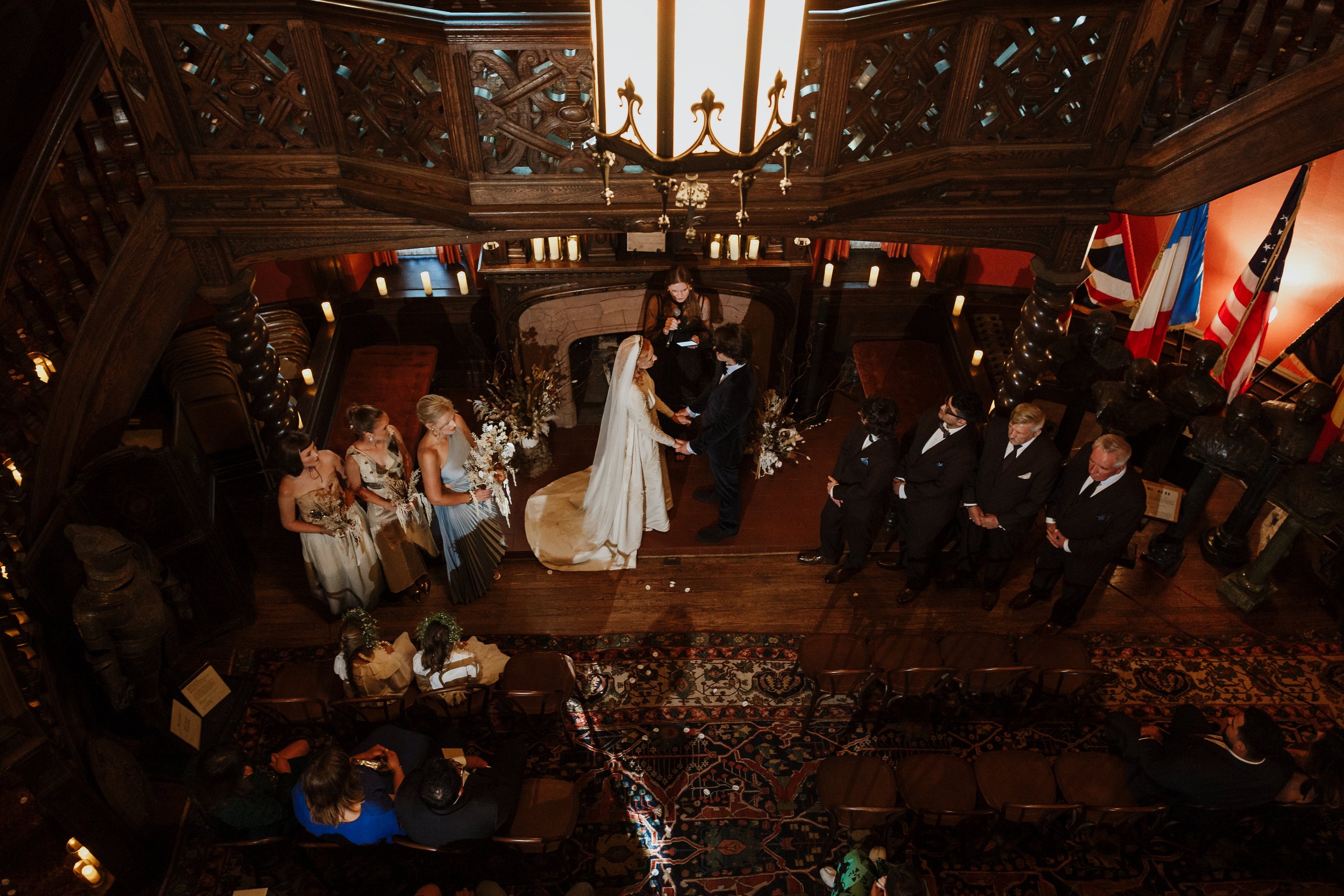 A wedding ceremony in an ornate, wood-paneled room with a high ceiling. The bride and groom stand at the altar, holding hands, with an officiant behind them. Bridesmaids and groomsmen are lined up on either side. The room is decorated with floral arr