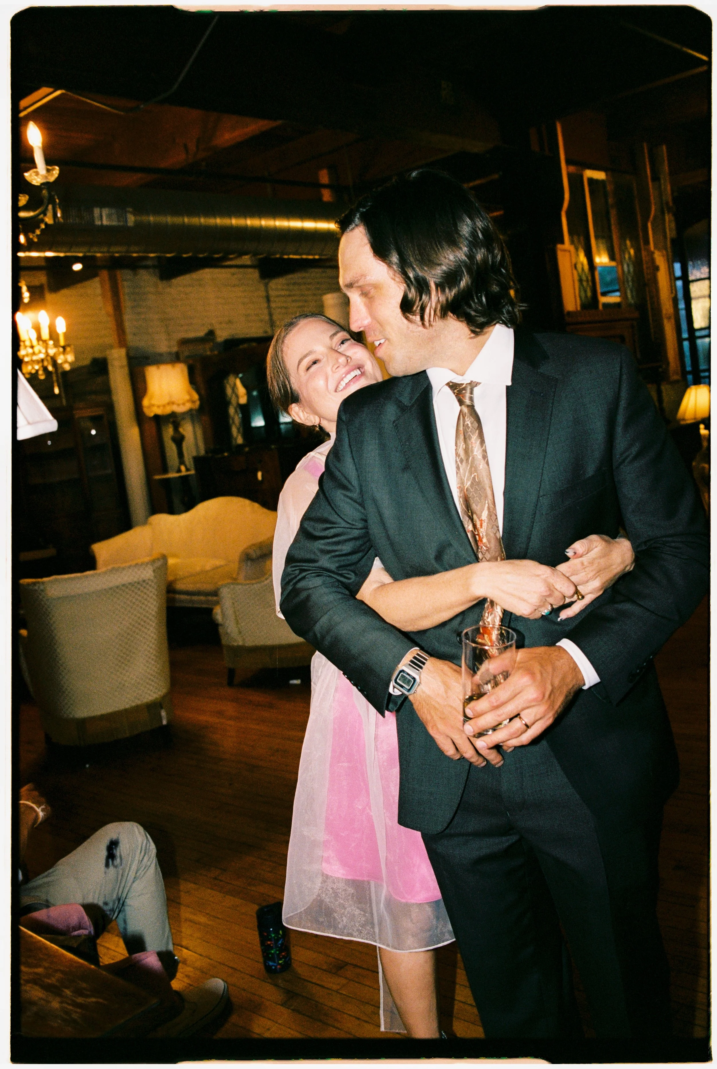 A man in a suit and a woman in a pink dress are embracing and smiling in a warmly lit restaurant or bar, holding a drink together.