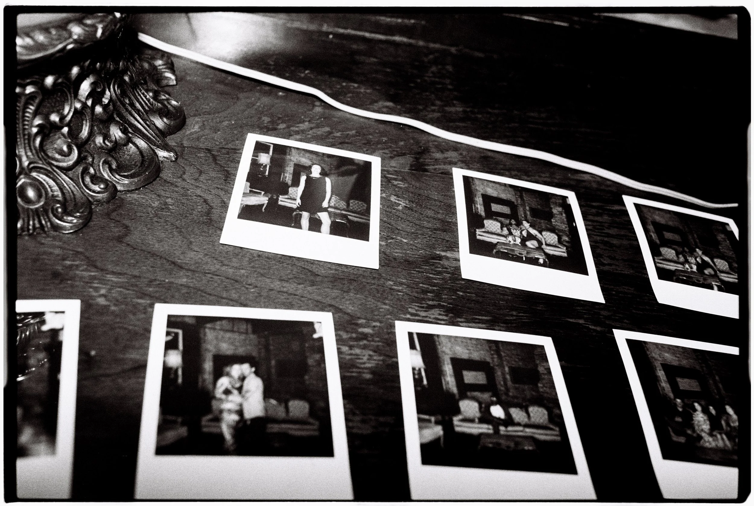 A collection of black and white printed photographs laid out on a wooden surface, with ornate decorative objects partially visible on the left side.