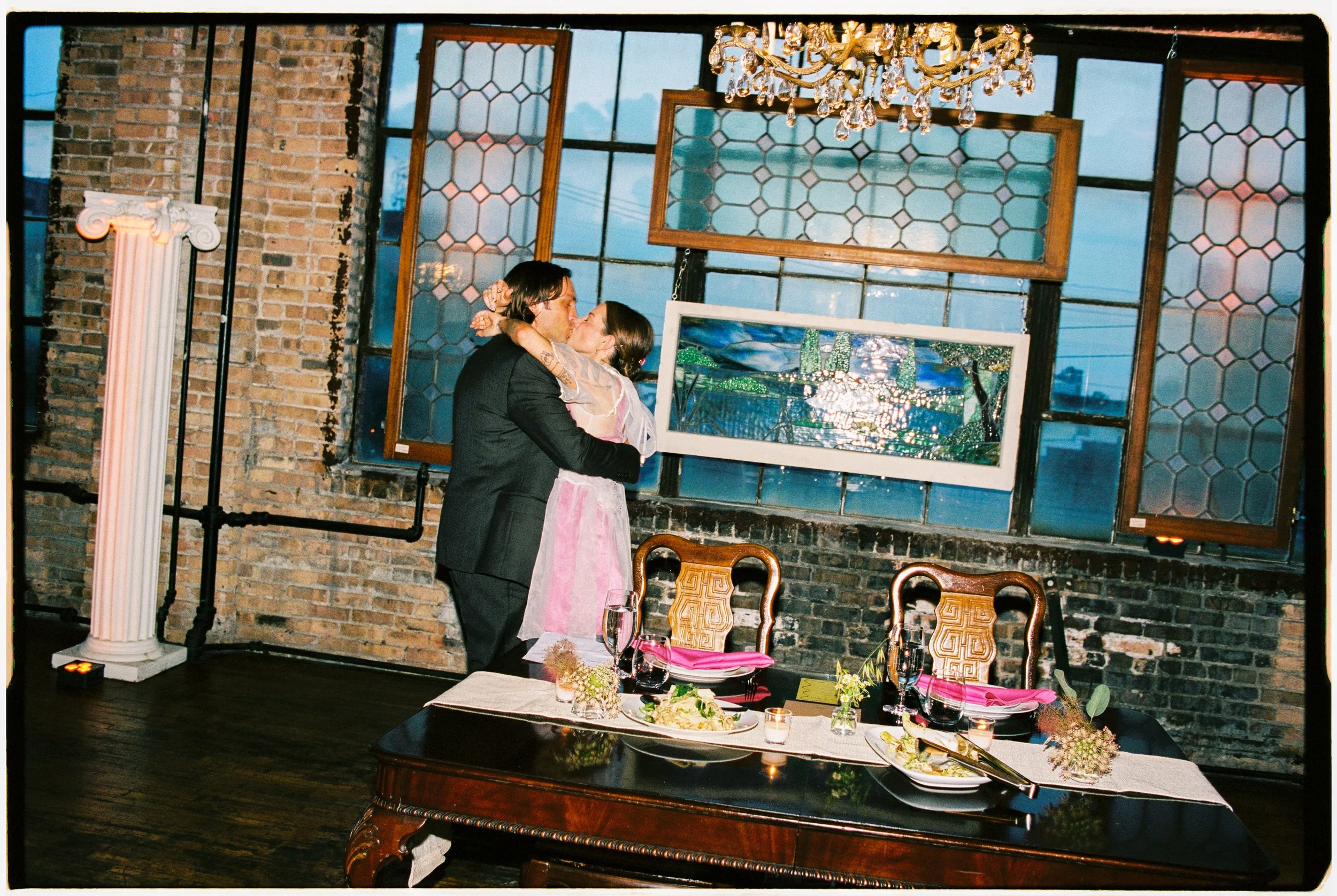 A couple sharing a kiss at a wedding reception. The woman wears a pink dress, and the man wears a black suit. The scene is set with a decorated table, floral arrangements, and candles, with a brick wall and stained glass windows in the background.
