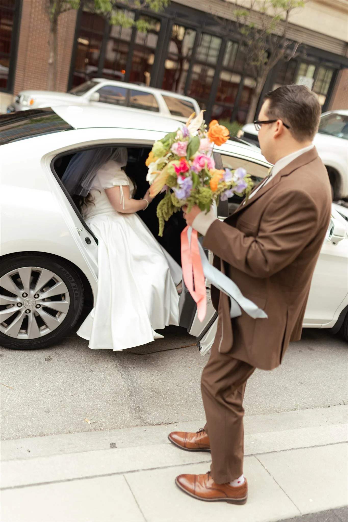 A man in a brown suit is handing a bouquet of flowers to a bride sitting in a white car. The bride is wearing a white dress and veil. The scene takes place outdoors on a city street.