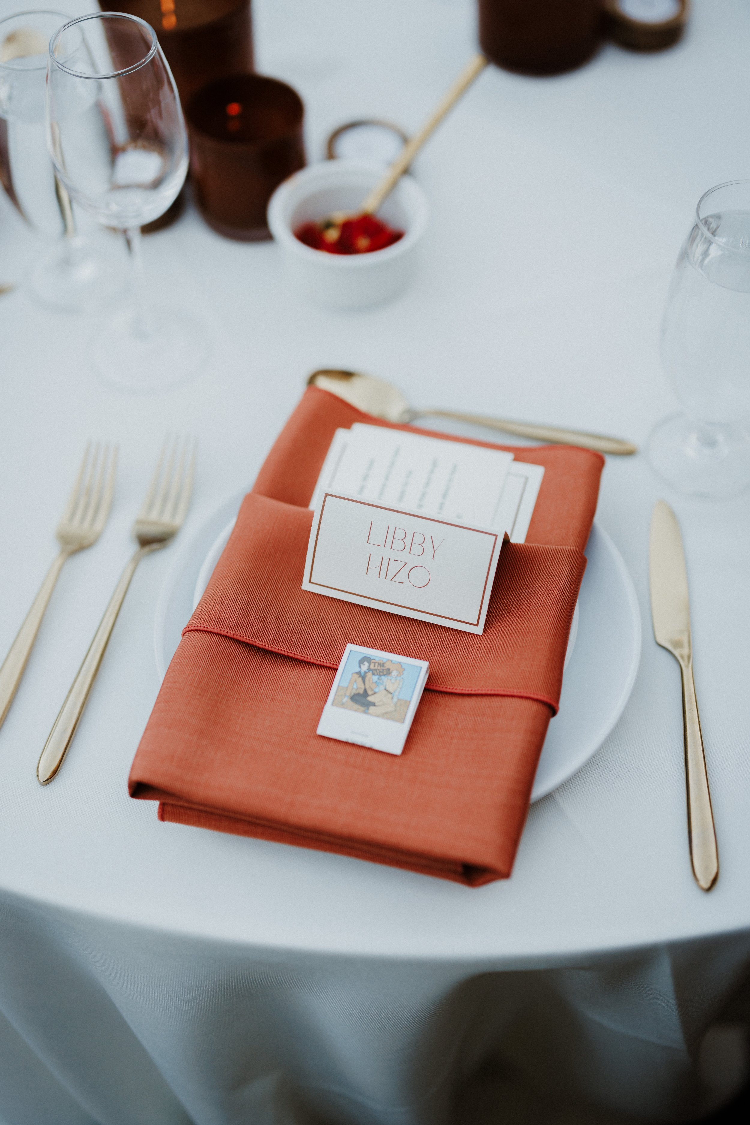A place setting on a white table with a folded coral napkin, a name card reading 'Libby Hizo', a small illustrated card, two forks, and two knives. In the background are clear and brown drinking glasses and a small white bowl with a red dish and a to