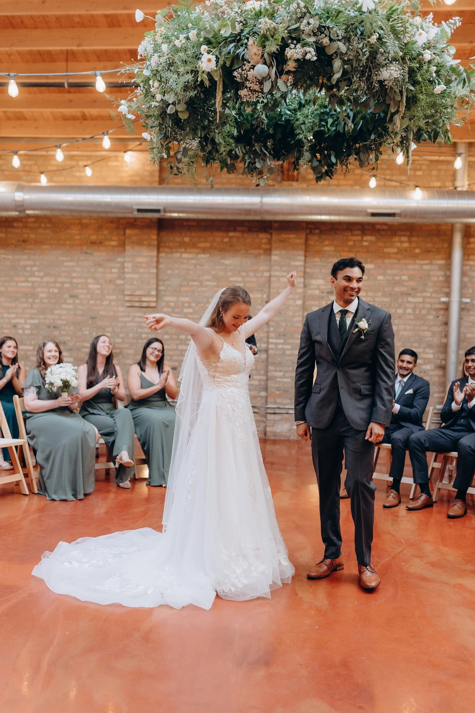 A bride and groom standing together during a wedding ceremony, with the bride raising her arms and the groom smiling, surrounded by guests seated on chairs in a rustic indoor venue with brick walls and string lights overhead.