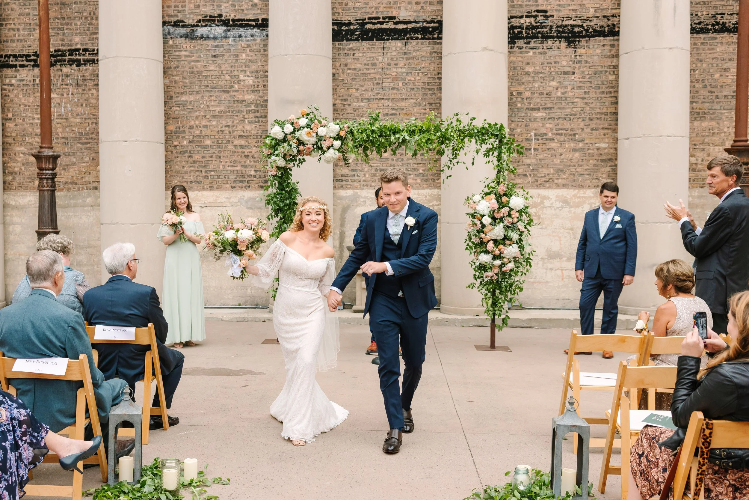 A bride and groom walking hand in hand down the aisle during their wedding ceremony in an outdoor urban setting with brick wall backdrop and floral arch. Guests seated on wooden chairs are watching and clapping, with some taking photos.