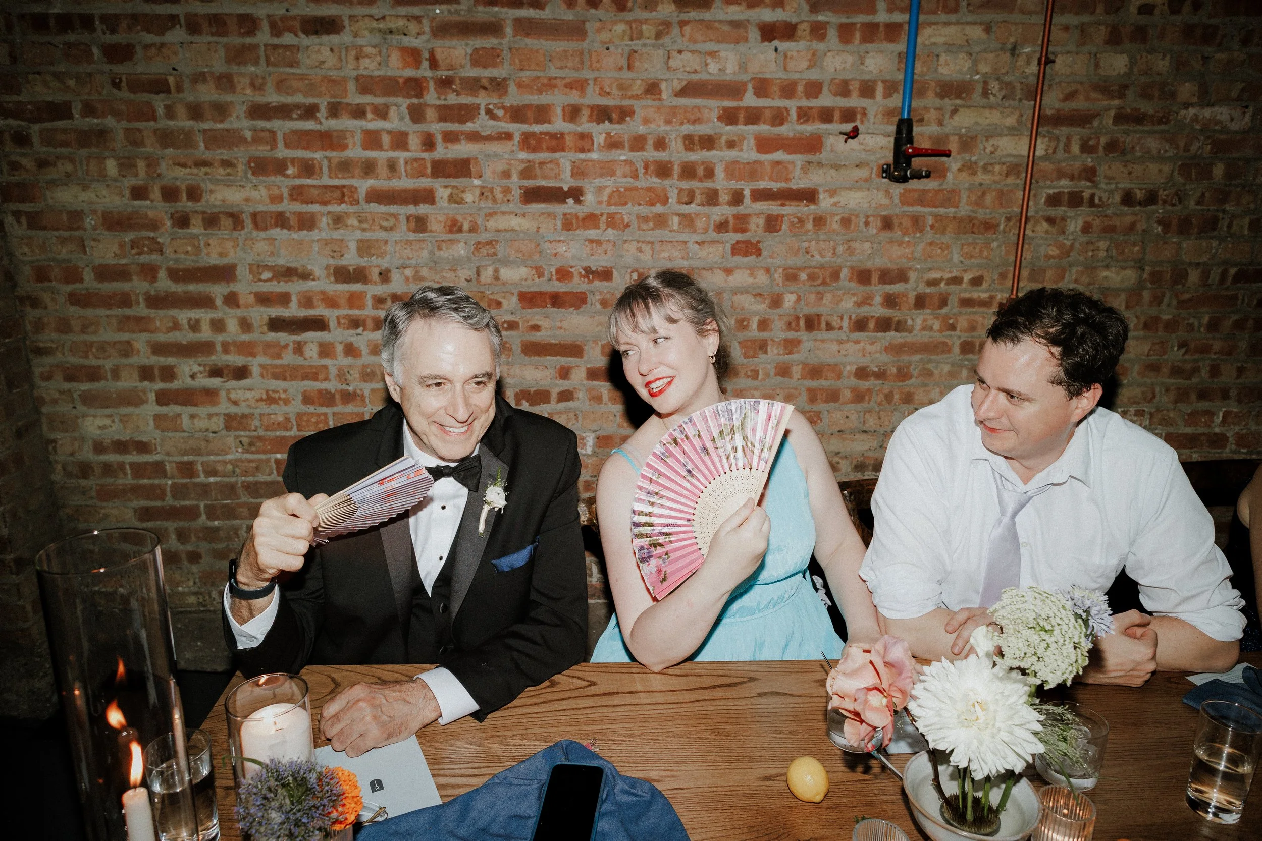 Three people sitting at a wooden table in front of a brick wall, two men and a woman dressed up for a formal event. The man on the left is wearing a black tuxedo and holding a paper fan, the woman in the middle is wearing a light blue dress and holdi