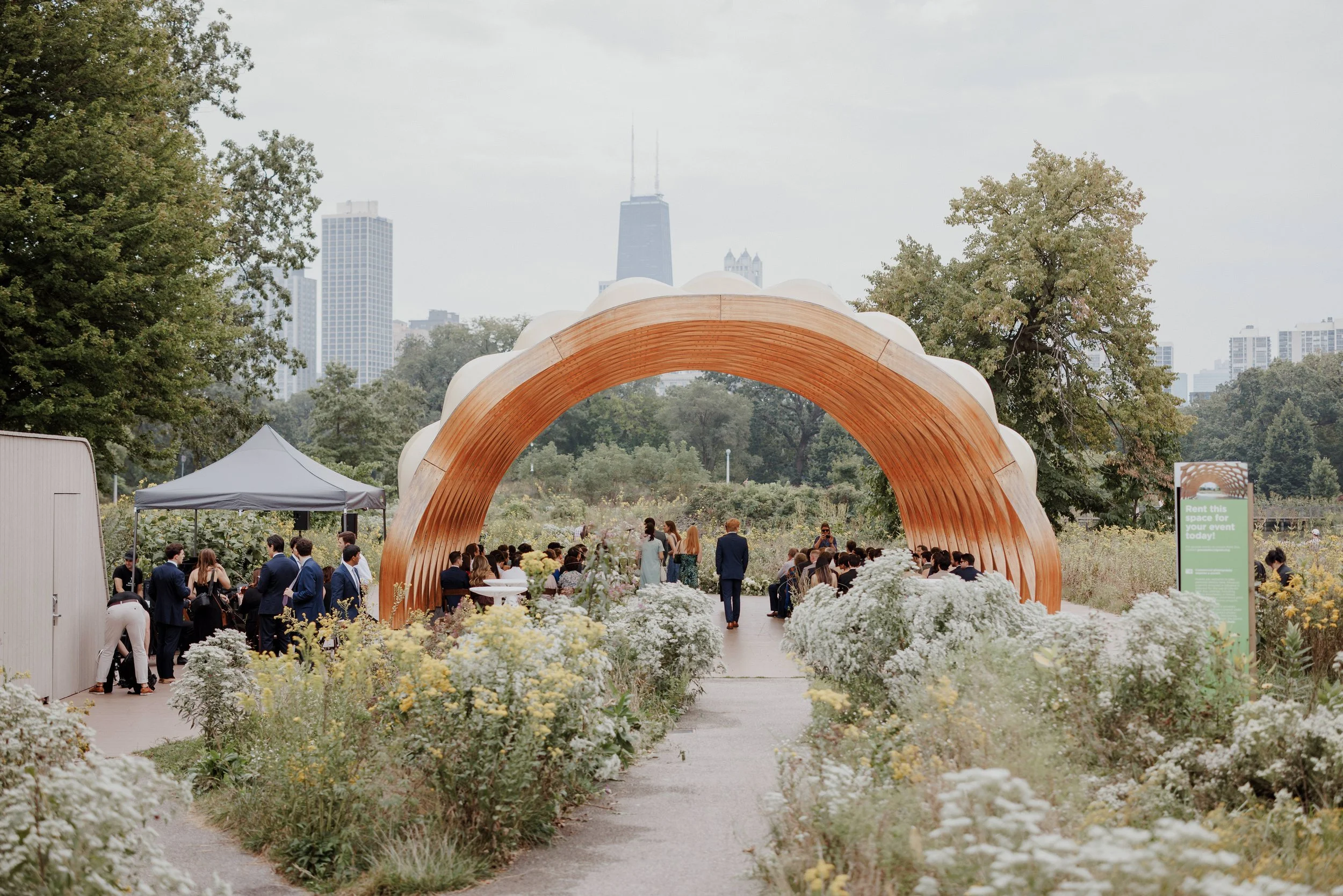 Outdoor wedding or event under a wooden archway with a city skyline, including the Willis Tower, in the background. There are people gathered, greenery, and white flowers along the pathway.