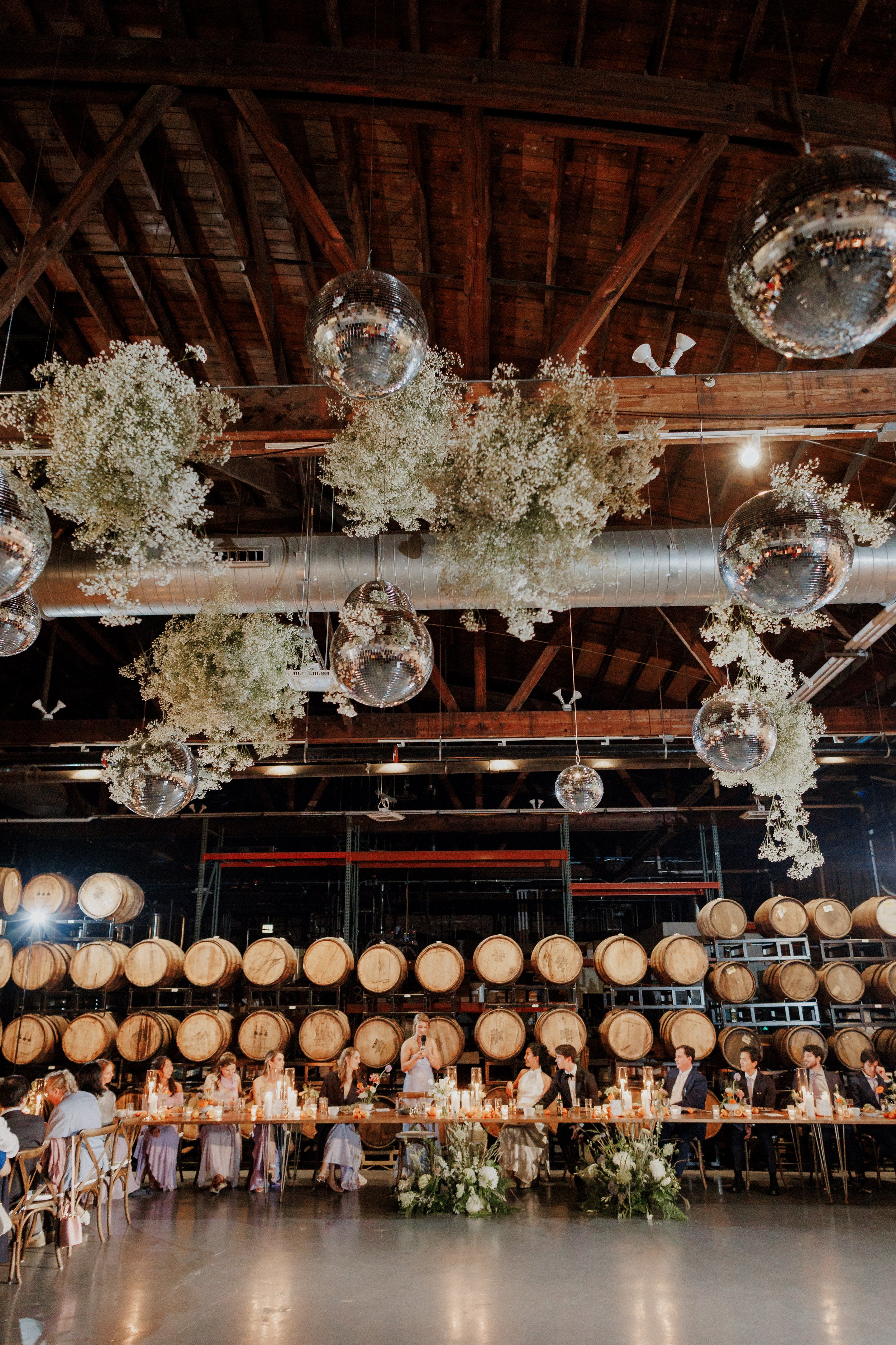 Wedding reception inside a rustic venue with wooden beams, hanging mirrors, surrounded by wine barrels, and a long table with guests.