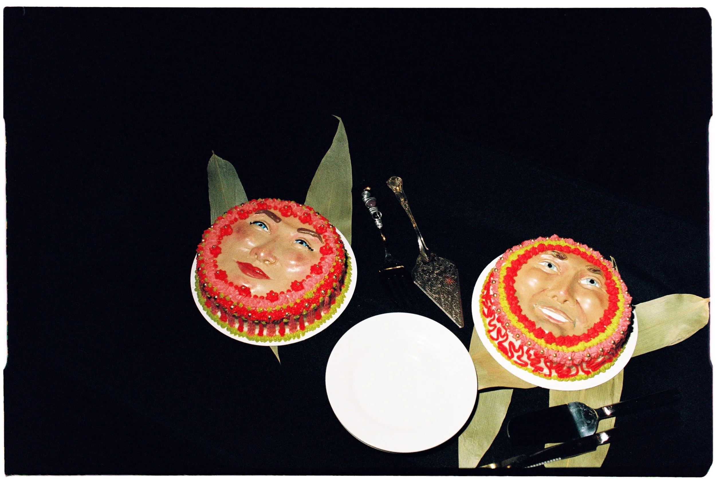Two decorative cakes with faces and red, pink, and yellow icing, surrounded by leaves, on a black table with empty white plates and utensils.