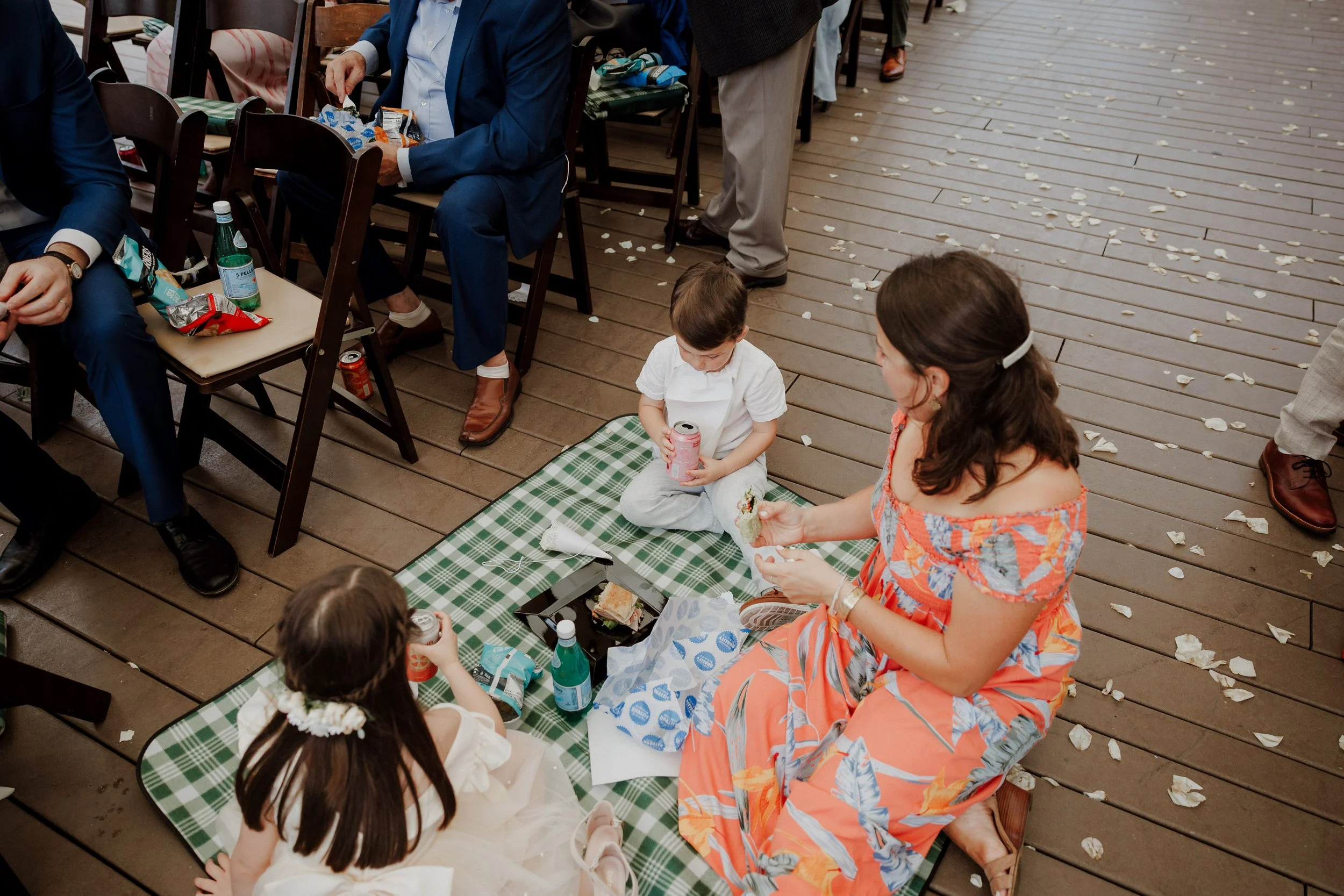 Children and adults sitting on a patterned picnic blanket on a wooden deck, surrounded by scattered flower petals, enjoying a meal and drinks.