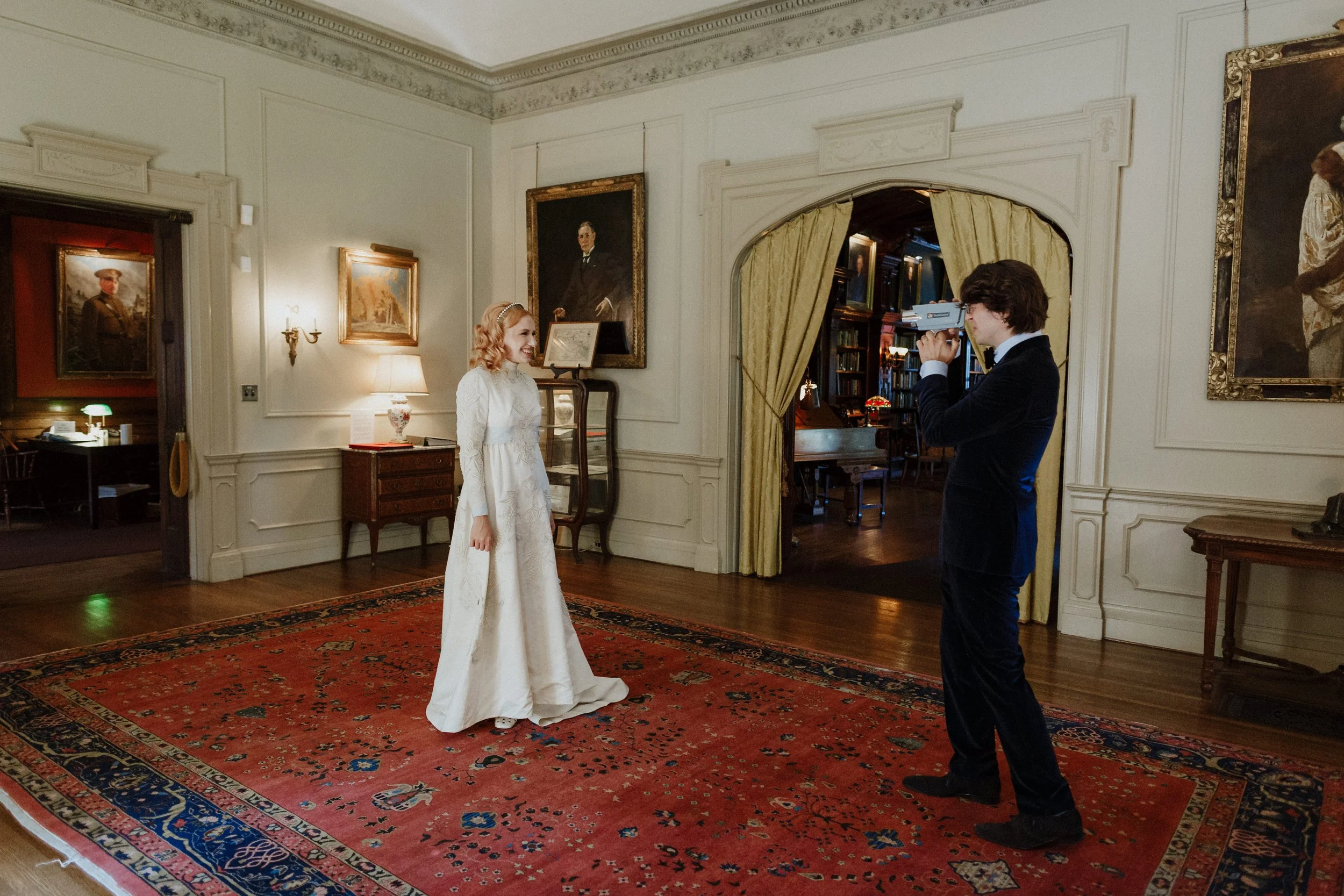 Young woman in a white vintage dress being photographed by a young man in a black suit inside a historic house or museum with portraits on the walls and a red patterned rug on the floor.