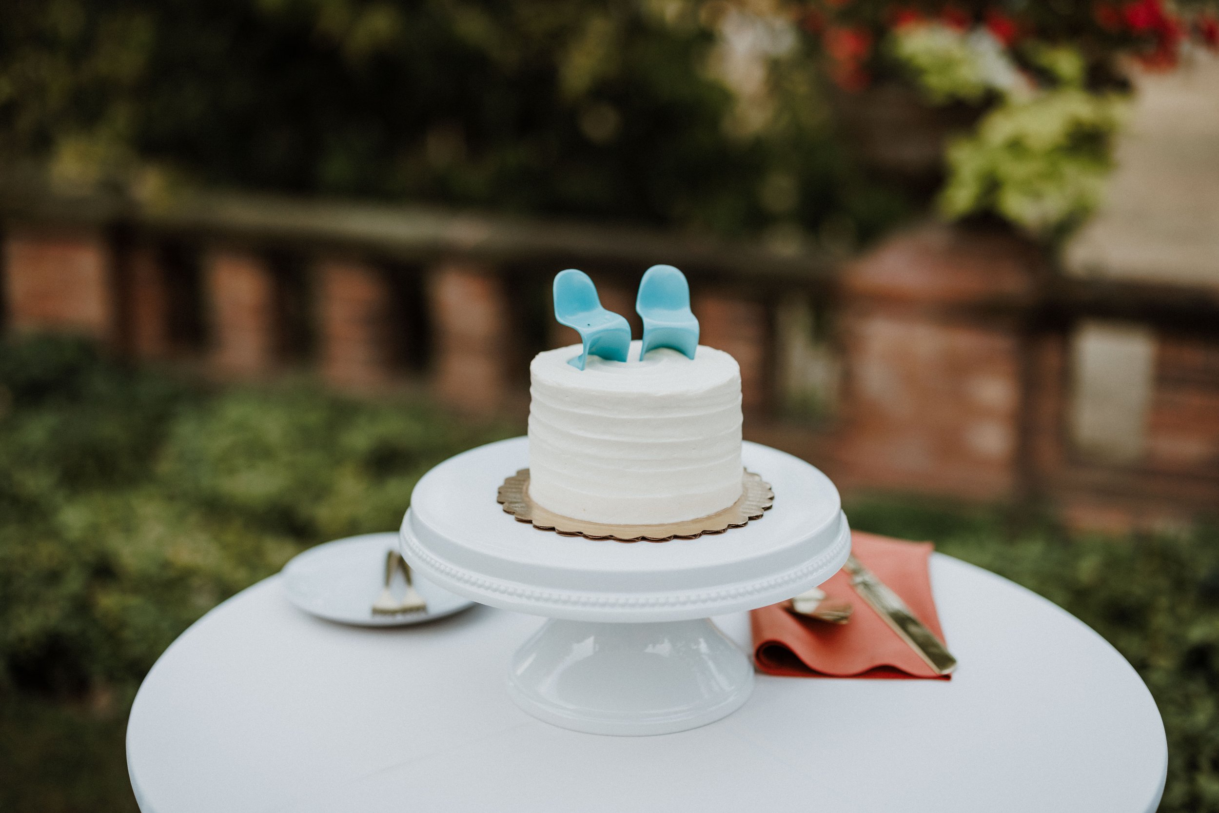 A small white cake with two blue plastic chairs on top, set on a white cake stand outdoors with green bushes and a brick wall in the background.