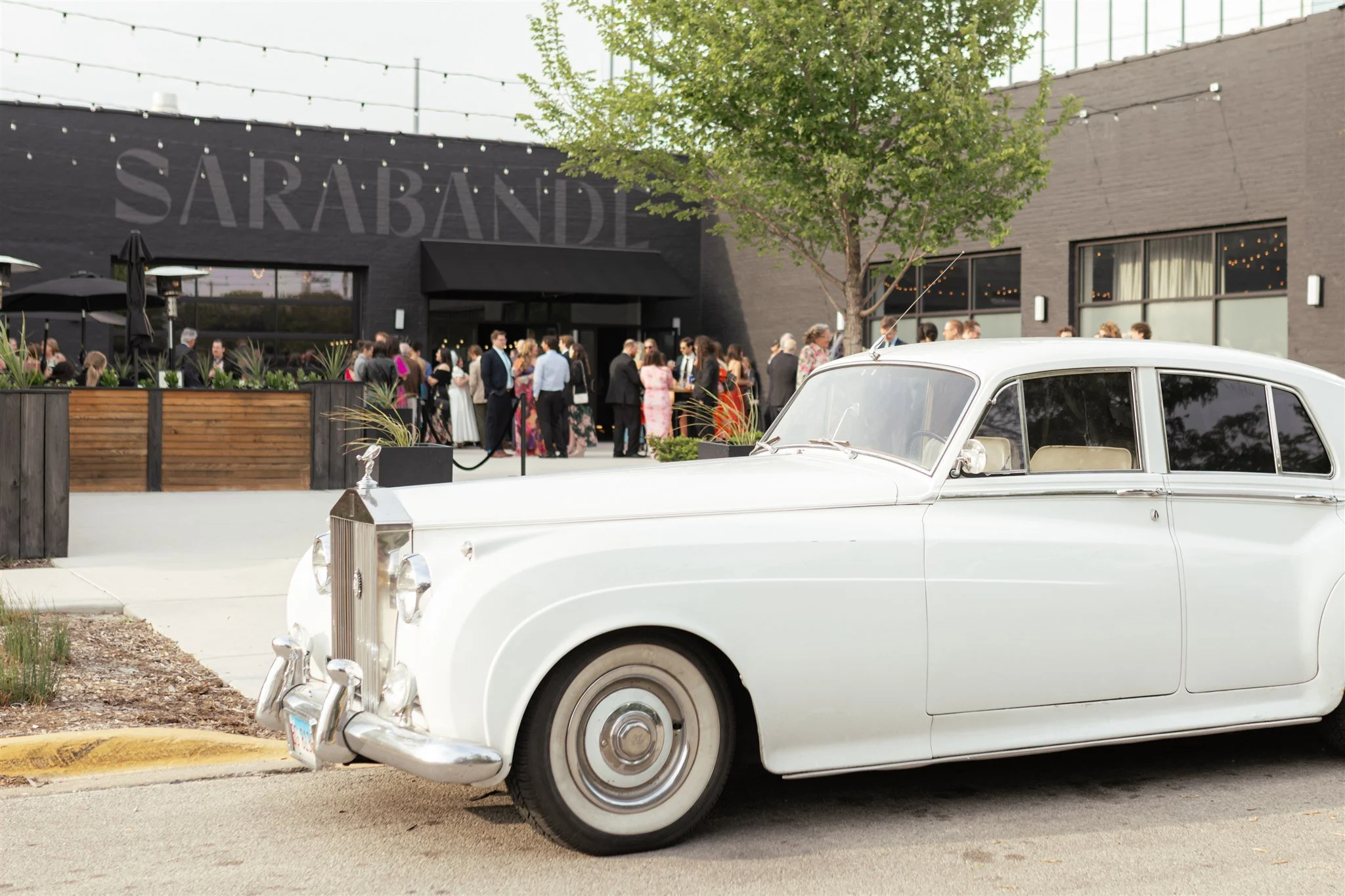 Vintage white car parked on the street in front of a building with a crowd of people gathered outside, and a tree providing some greenery.