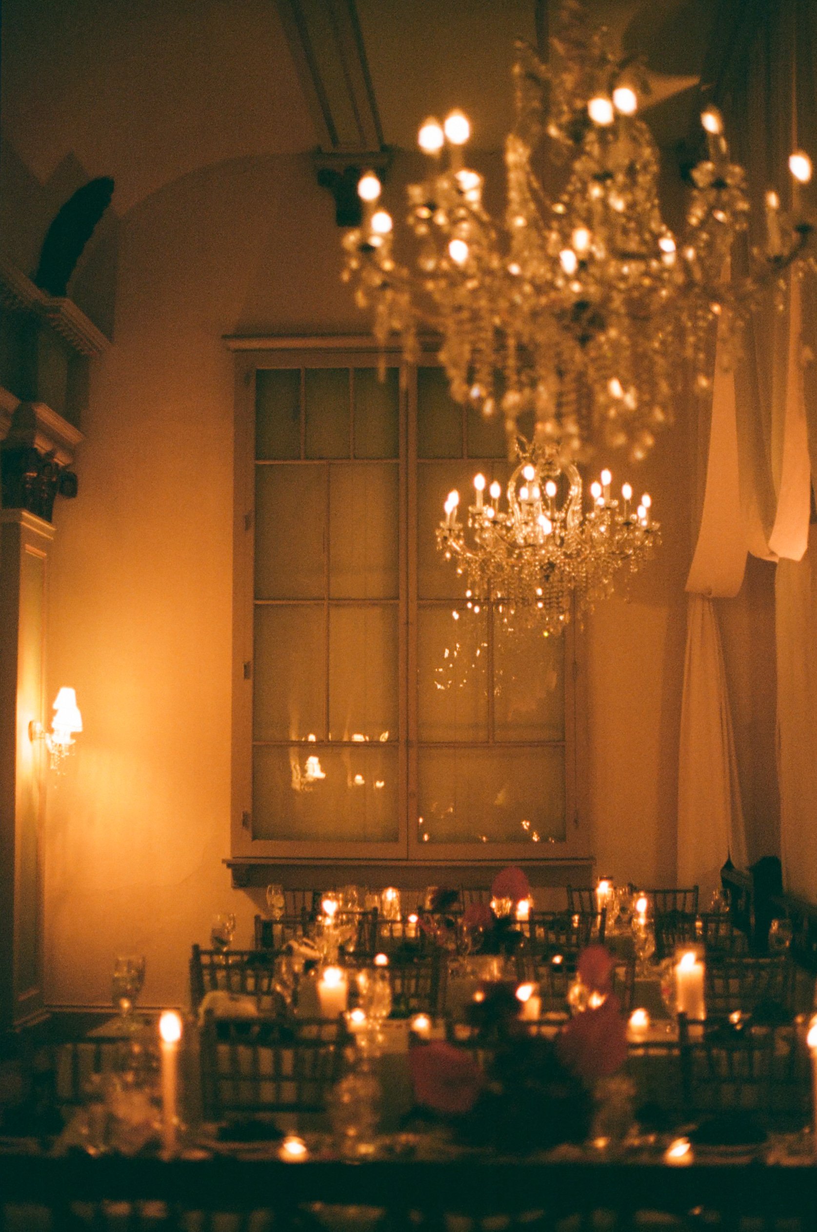 Elegant banquet hall with chandeliers, candlelit tables, and large window with curtains.