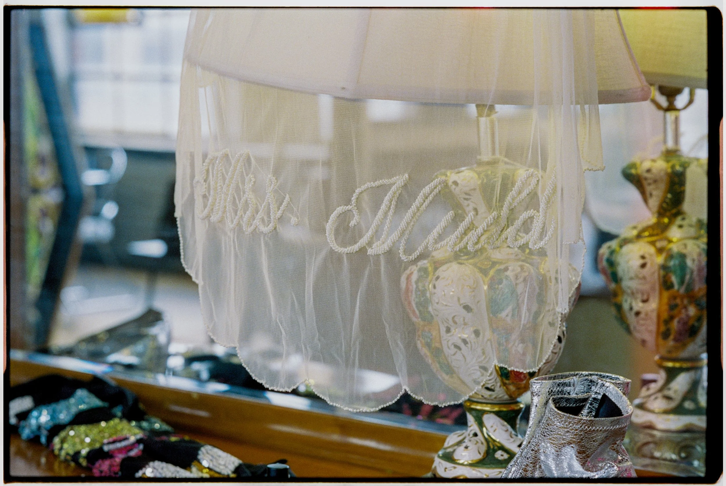 A display of vintage lace and embroidered fabric with the words 'Hand Embroidery' embroidered in cursive on a sheer fabric. Decorative ceramics and jewelry are visible in the background, indicating a craft or antique shop setting.