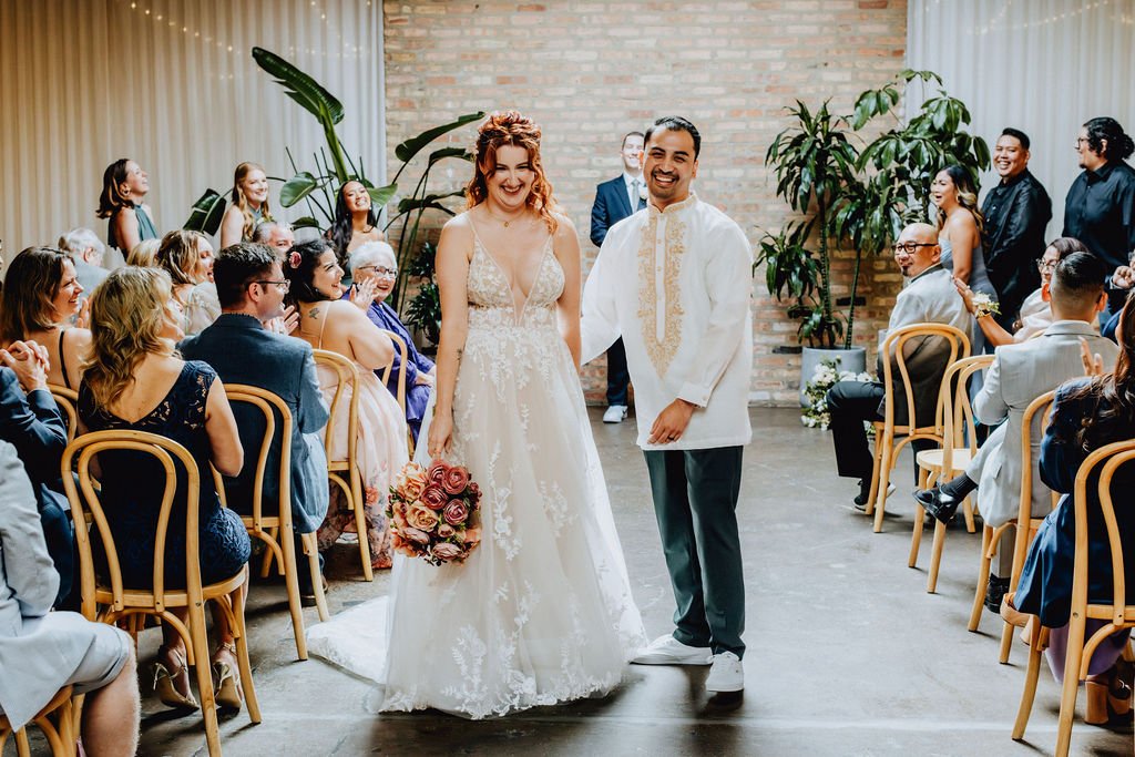 A bride and groom stand hand in hand at their wedding ceremony, smiling, surrounded by seated guests in a decorated indoor venue with plants.