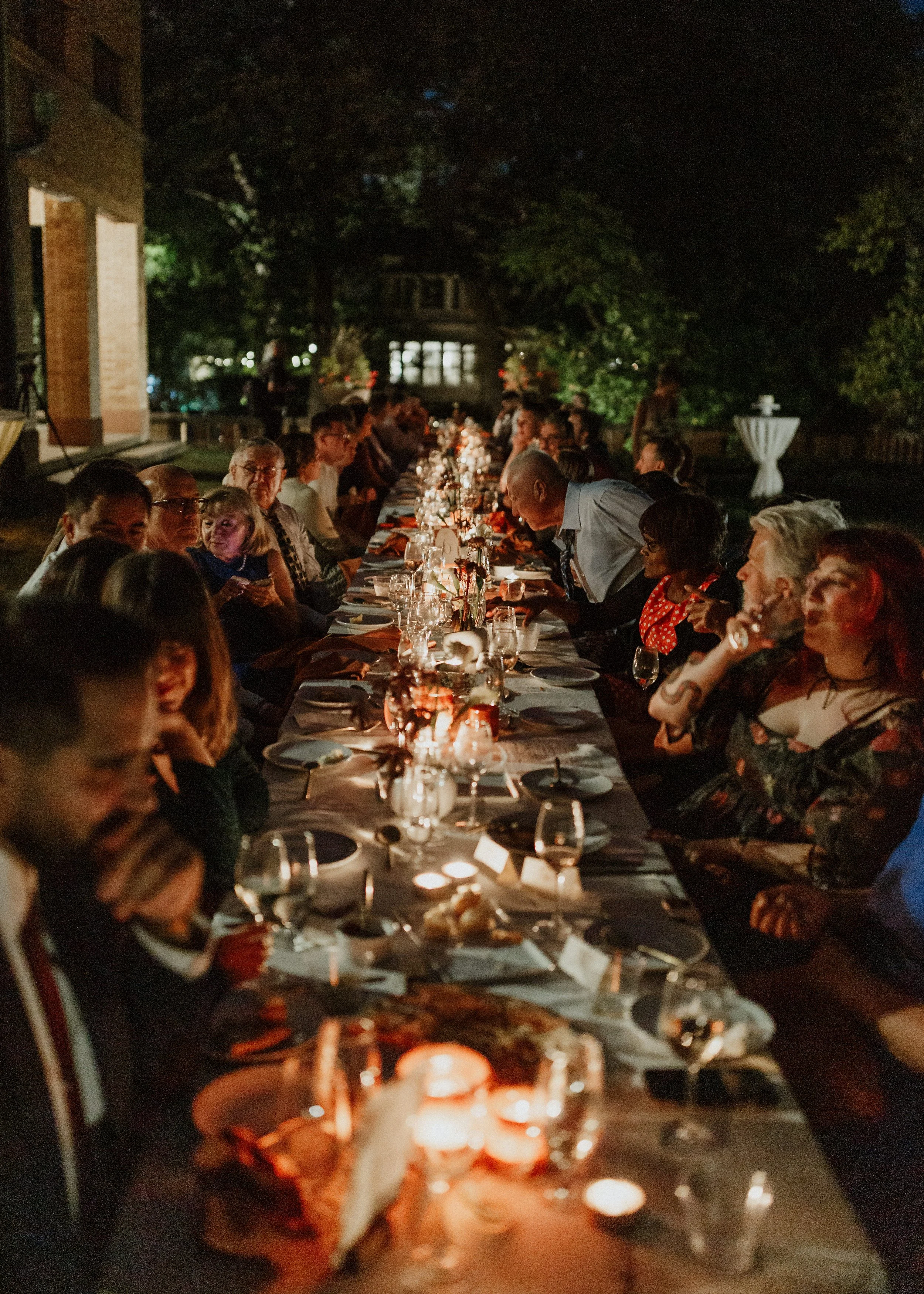 People gathered around a long dinner table outdoors at night, with candles and floral centerpieces, engaging in conversation and enjoying a formal dinner.