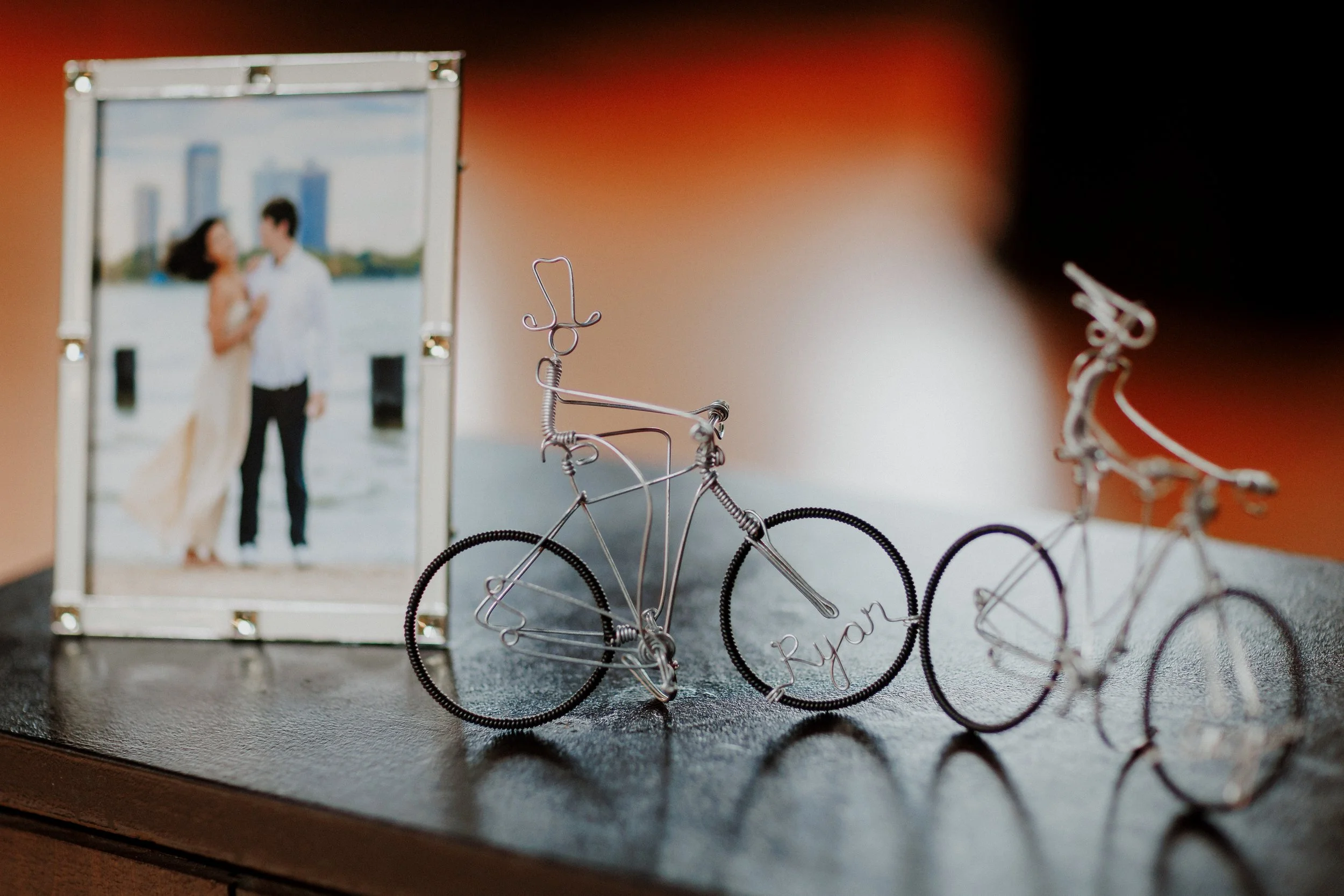 Small wire bicycle-shaped jewelry holder with a heart-shaped top, displaying a name, placed on a surface next to a blurred photo of a couple embracing outdoors.