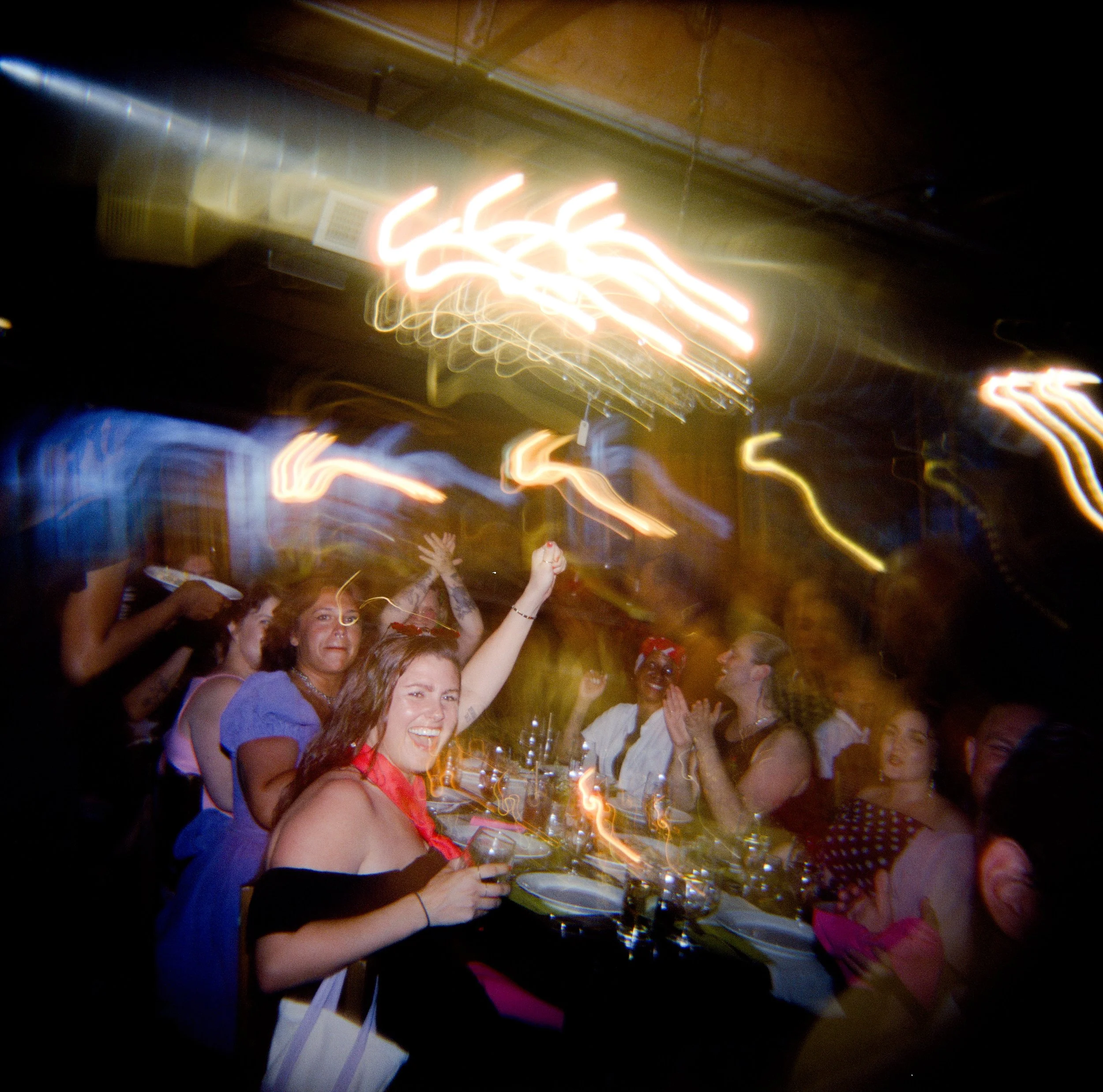 People celebrating at a party with some holding up glasses, in a dimly lit room with colorful lights.