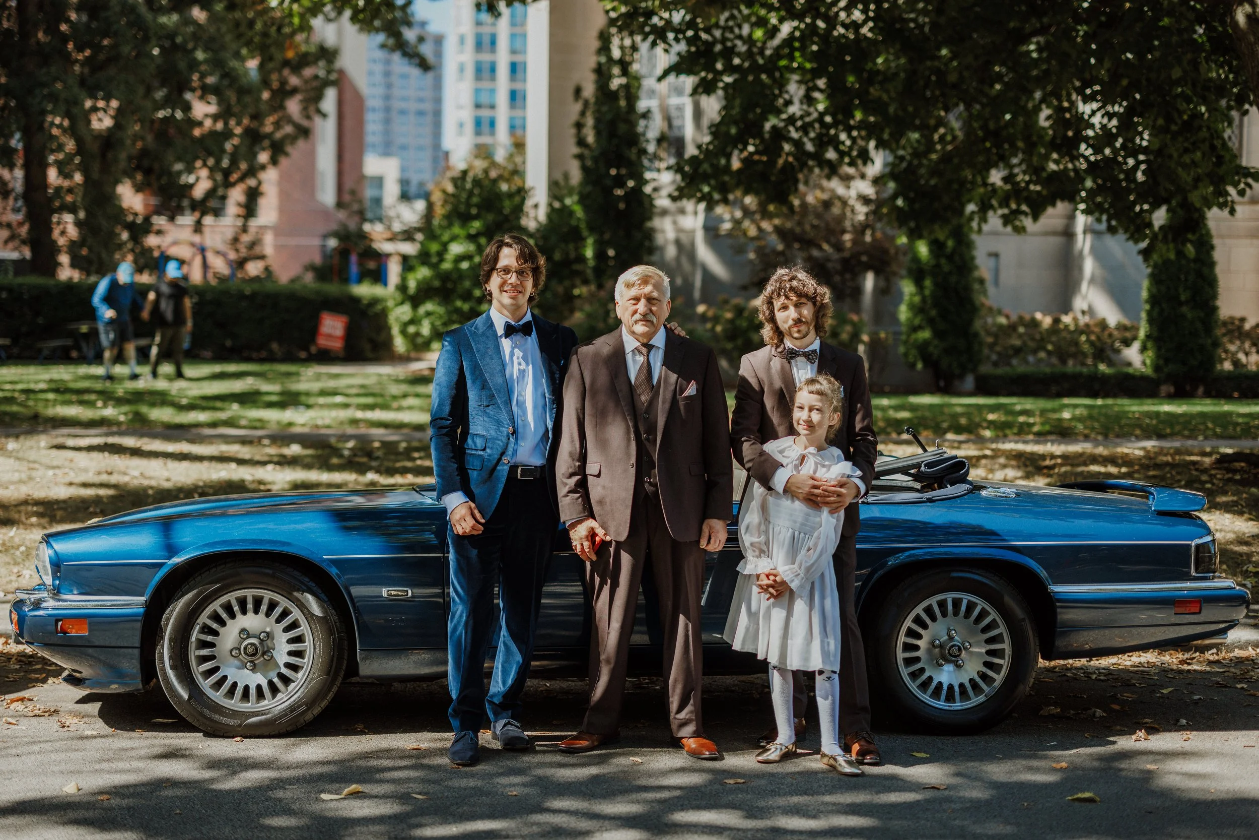 Four people standing in front of a blue classic car in a park, with trees and tall buildings in the background. The group includes two men and a young girl, all dressed in formal attire.