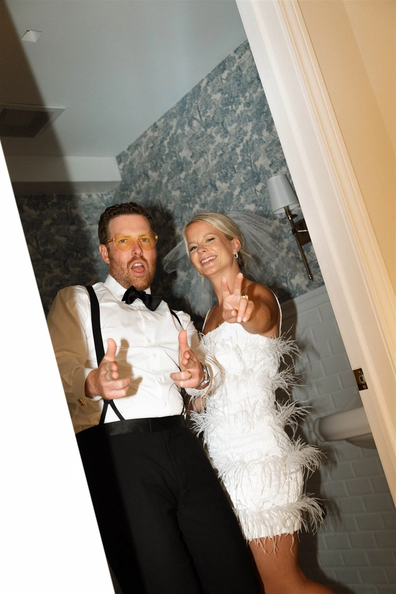 A man in formal attire with a white shirt, black bowtie, and suspenders, and a woman in a white feathered dress and veil, both making playful gestures and smiling at the camera in a room with blue patterned wallpaper and a beige wall.